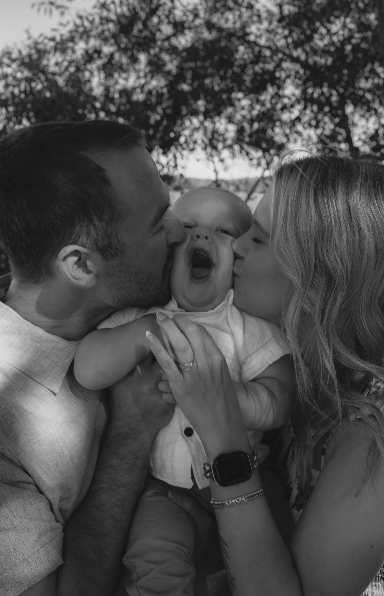 A series of black and white photos showing a couple sharing tender kisses while holding a baby between them outdoors.