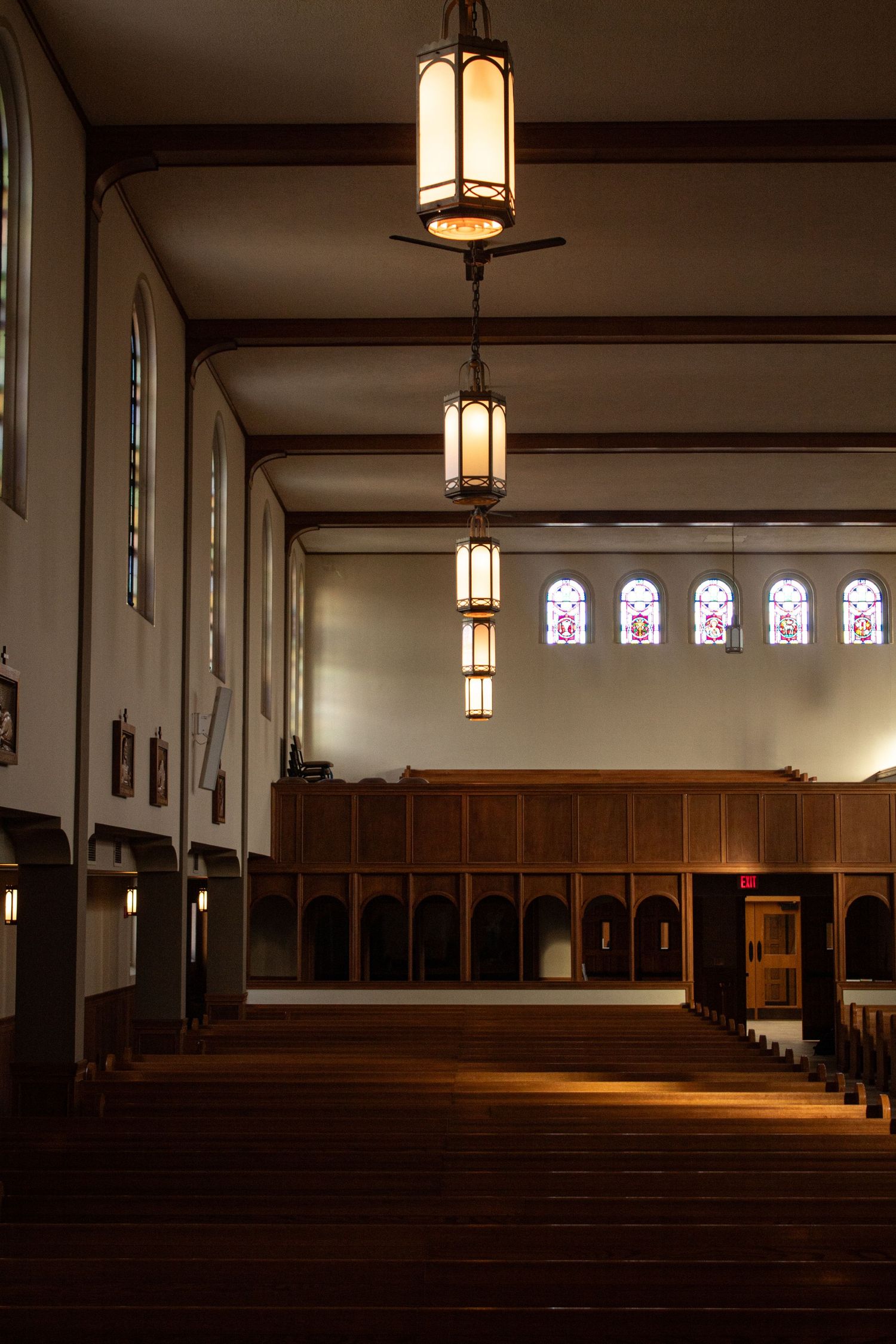 Interior of empty church sanctuary with wooden pews, pendant lights, and stained glass windows along one side.