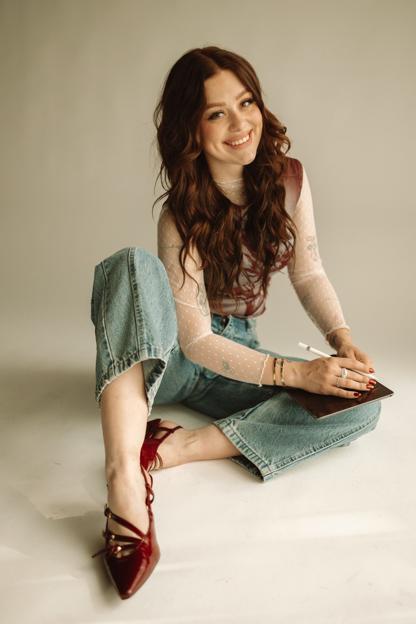 Woman in a pink sweater and denim skirt lounges on a white floor with red shoes while reading.