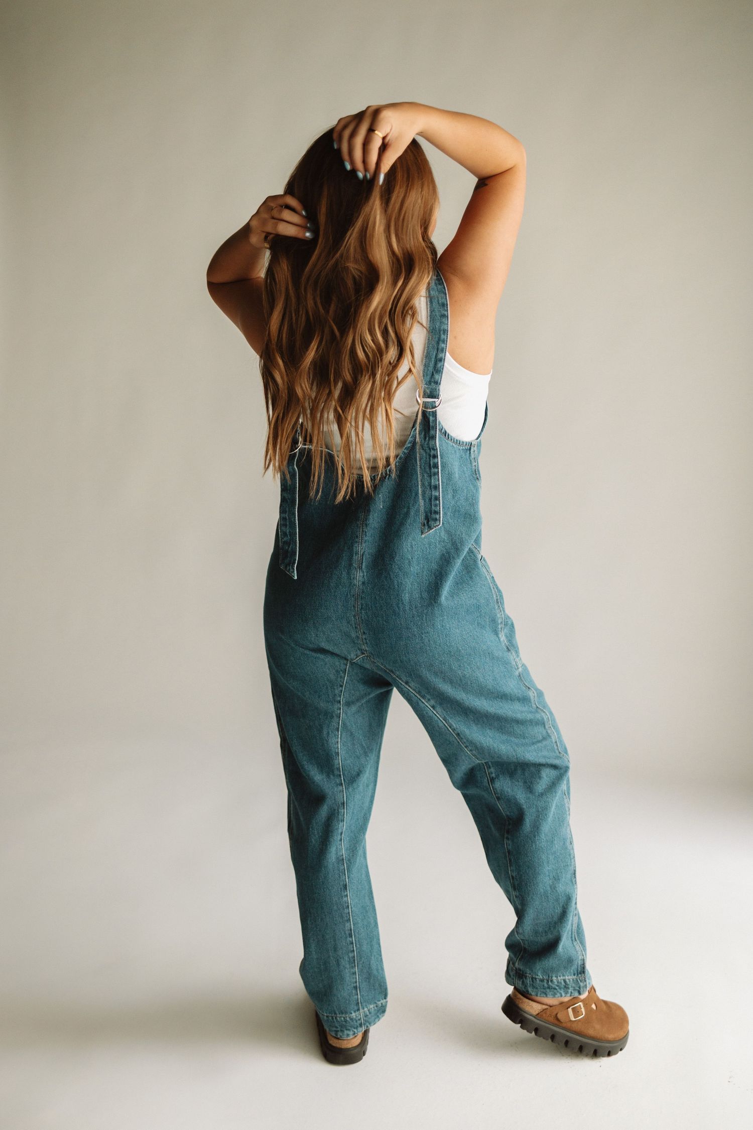 A person wearing denim overalls and brown boots stands backwards, running fingers through long wavy brown hair.