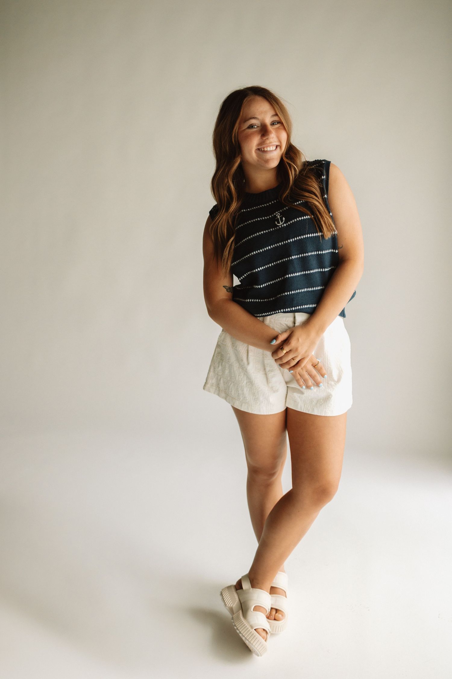 Woman in striped sleeveless top and white shorts poses against light backdrop with platform sandals.