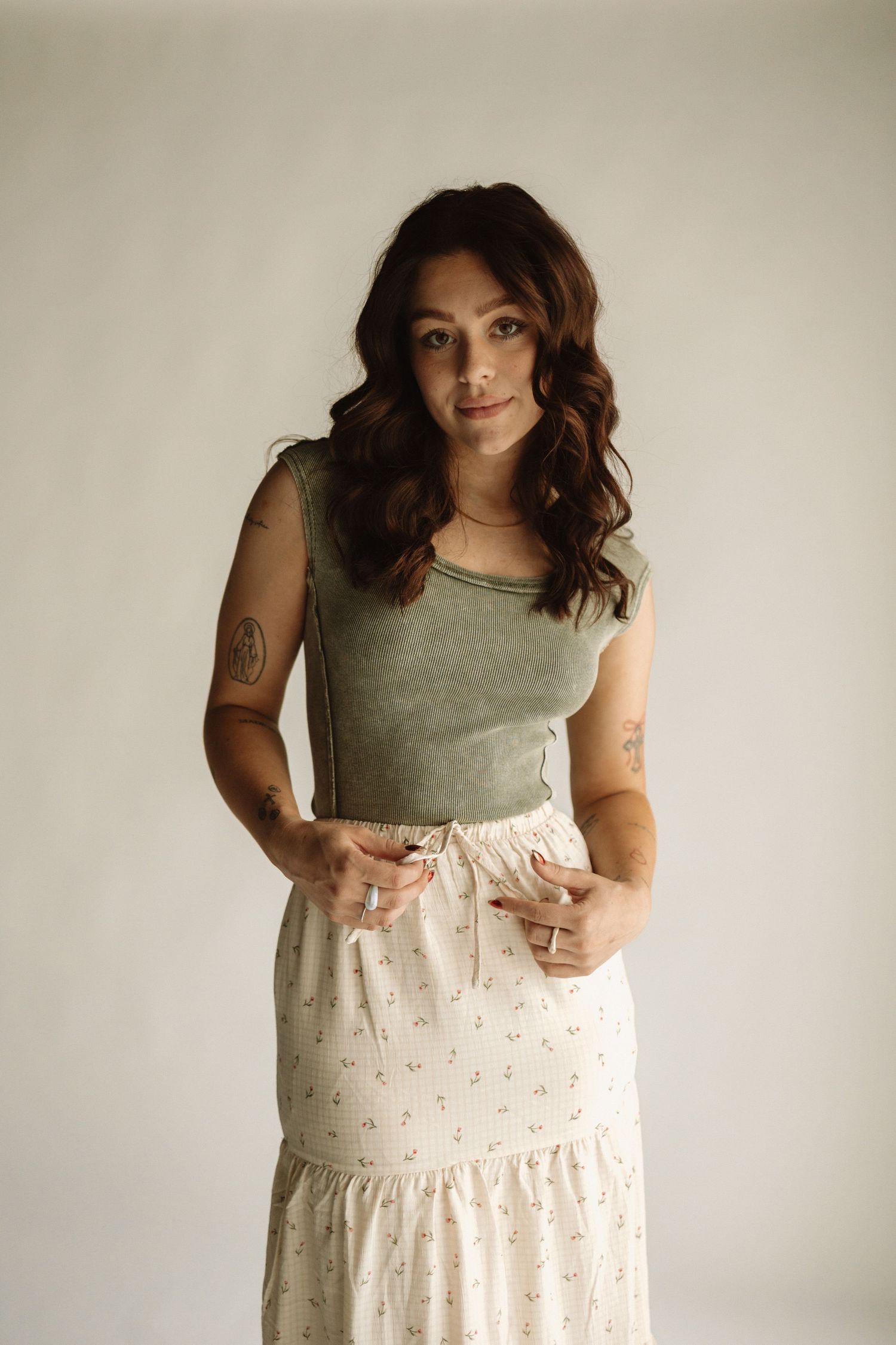 Person wearing olive sleeveless top and long cream-colored floral skirt in a minimalist photoshoot against light wall.