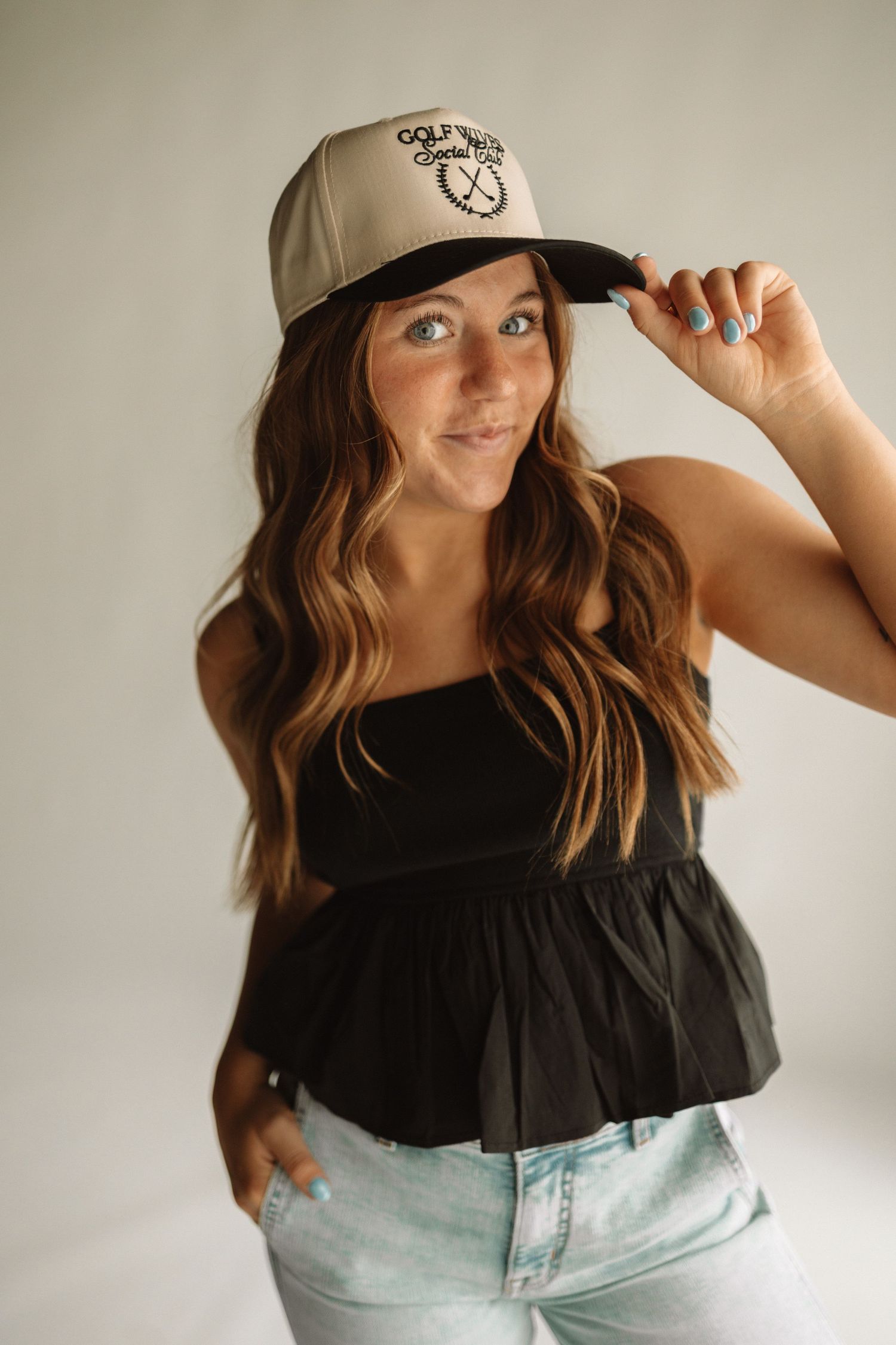 Woman wearing a black t-shirt and tan baseball cap poses playfully in front of white wall.