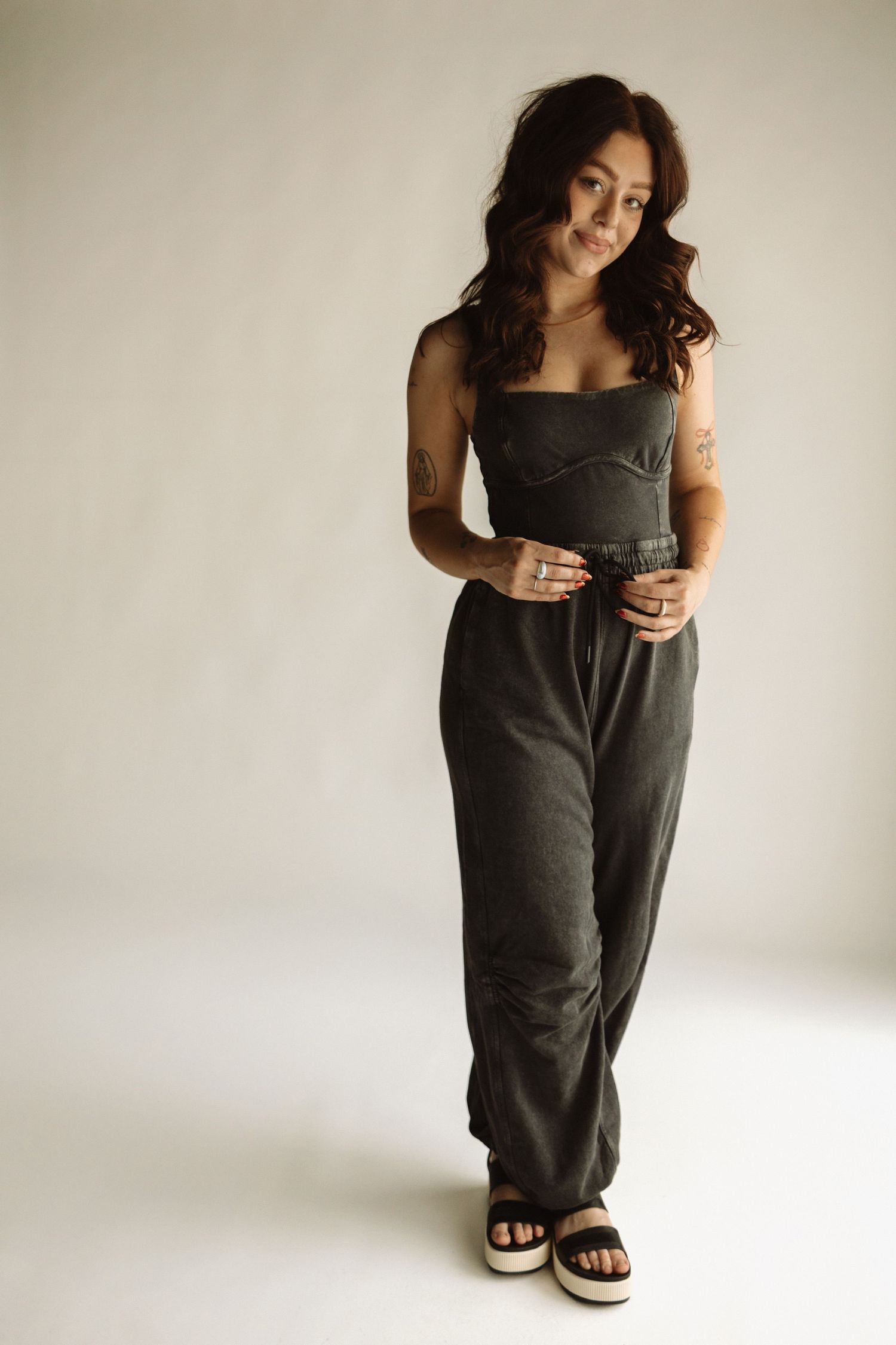 A person in a dark sleeveless jumpsuit and platform sandals poses against a light background in a studio setting.