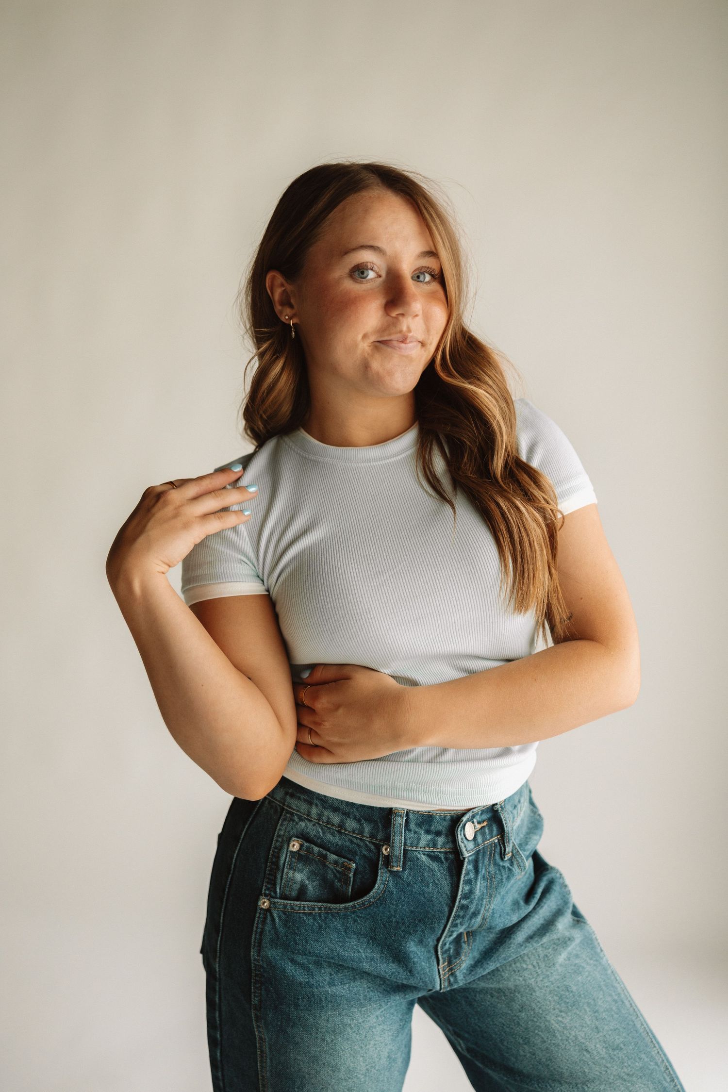 Standing casually against a white wall wearing a cropped white t-shirt and high-waisted blue jeans.