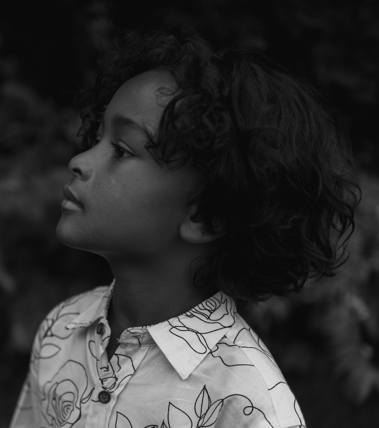 Person wearing white collared outfit looks upward with curly hair in dramatic black and white portrait composition.