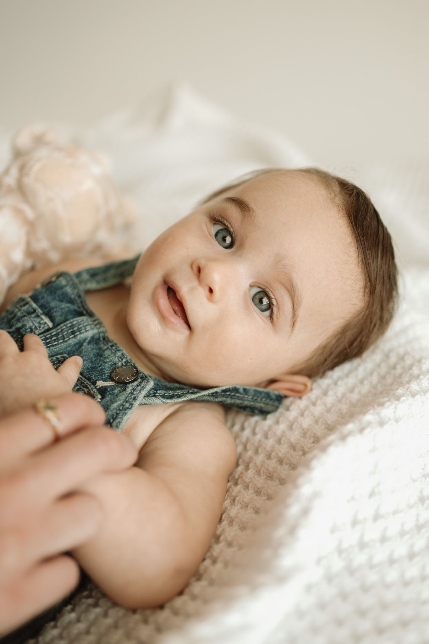 Sweet baby in teal outfit lying on white knitted blanket with soft lighting and dreamy expression.