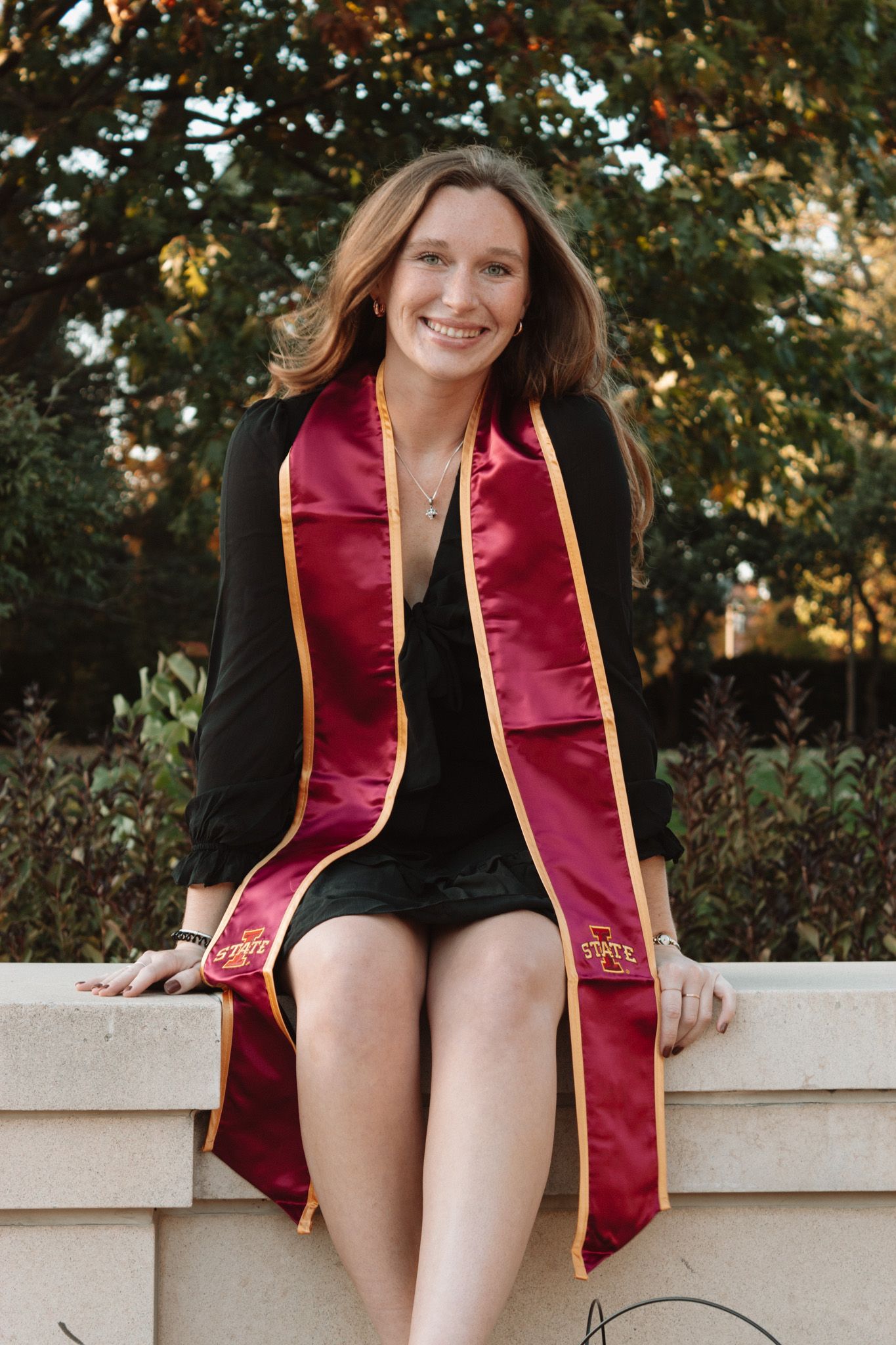 Graduate wearing maroon and gold stole with black dress sits on stone wall during outdoor graduation photoshoot.