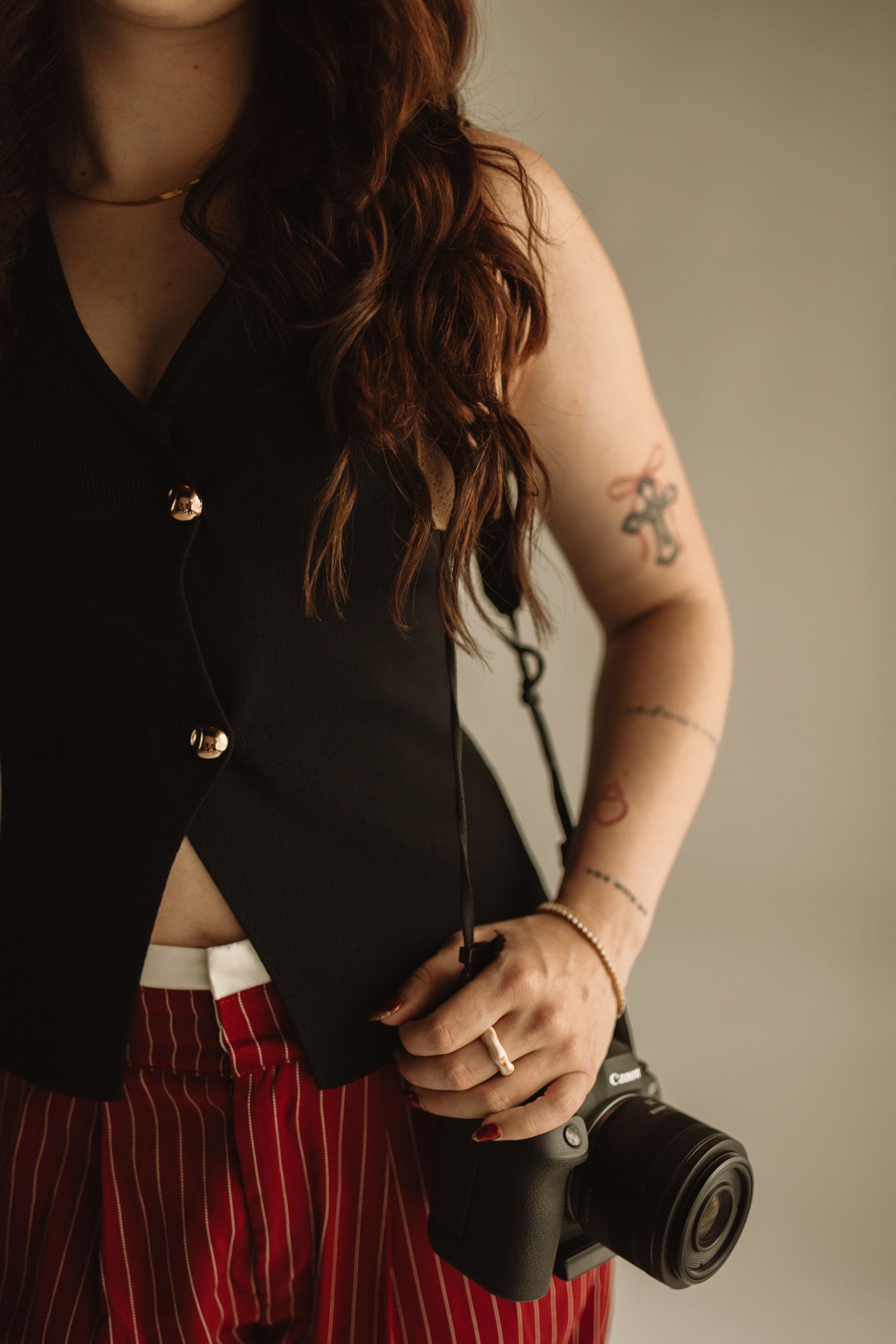 Professional photographer wearing black top and red striped pants holds camera with visible arm tattoos in studio setting.