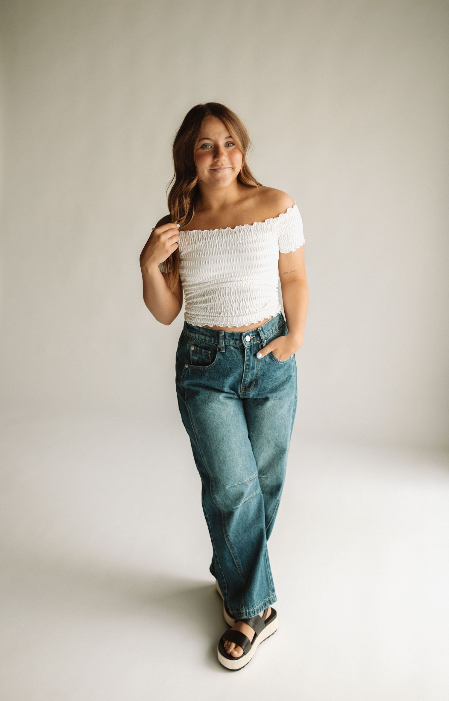 A model wearing a white off-shoulder smocked top, baggy blue jeans and platform sandals poses against a light background.