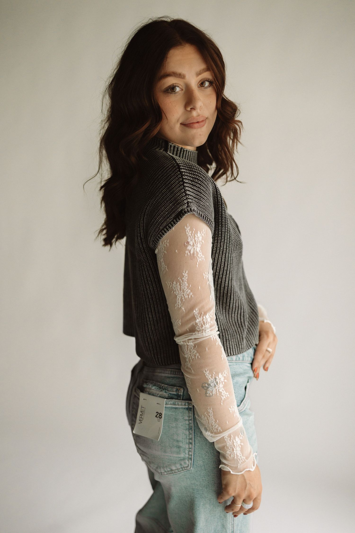 Model wearing grey knit vest with delicate white lace sleeves and light wash denim jeans against neutral background.