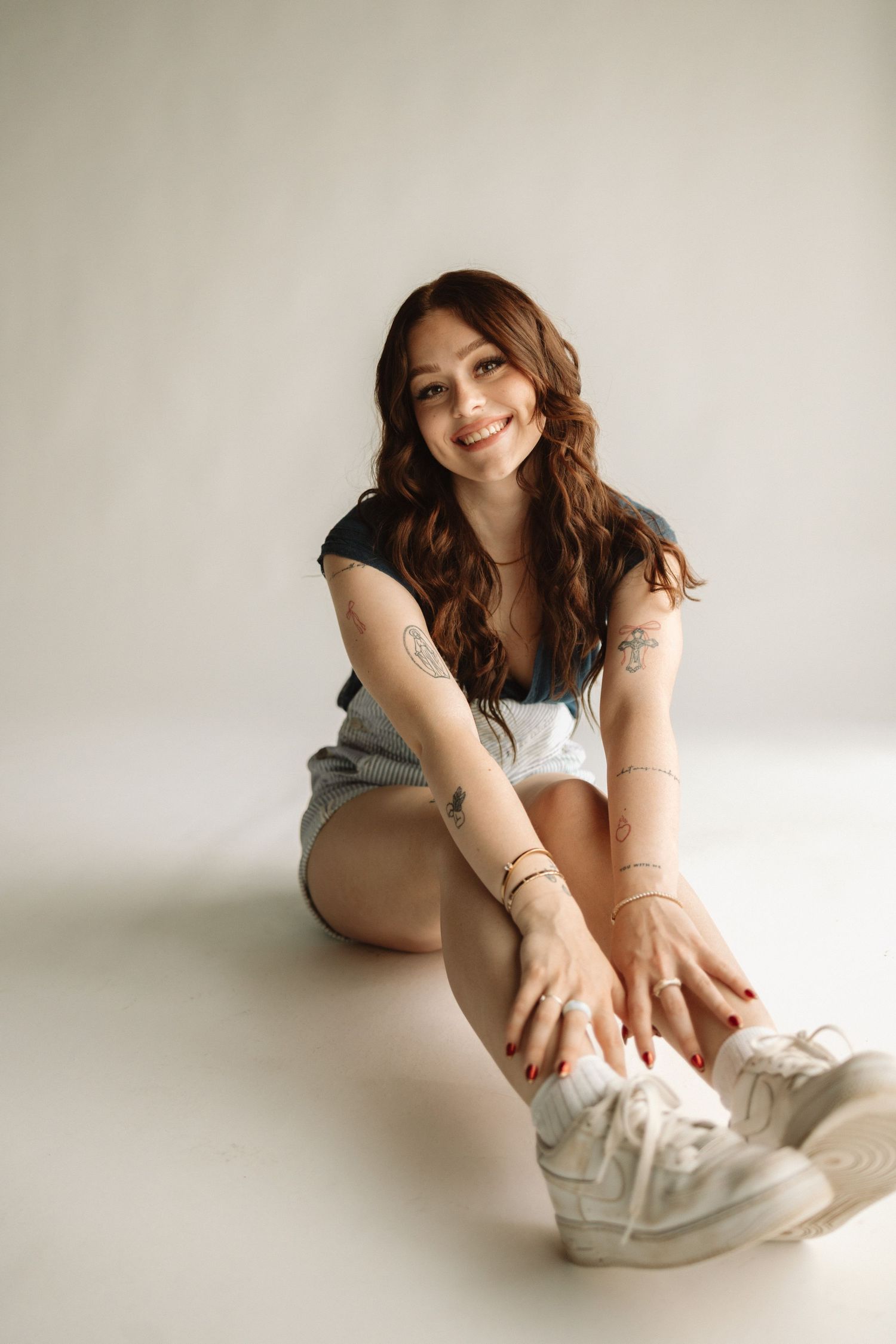 Young woman in casual attire sitting on floor wearing white sneakers and smiling against neutral background.