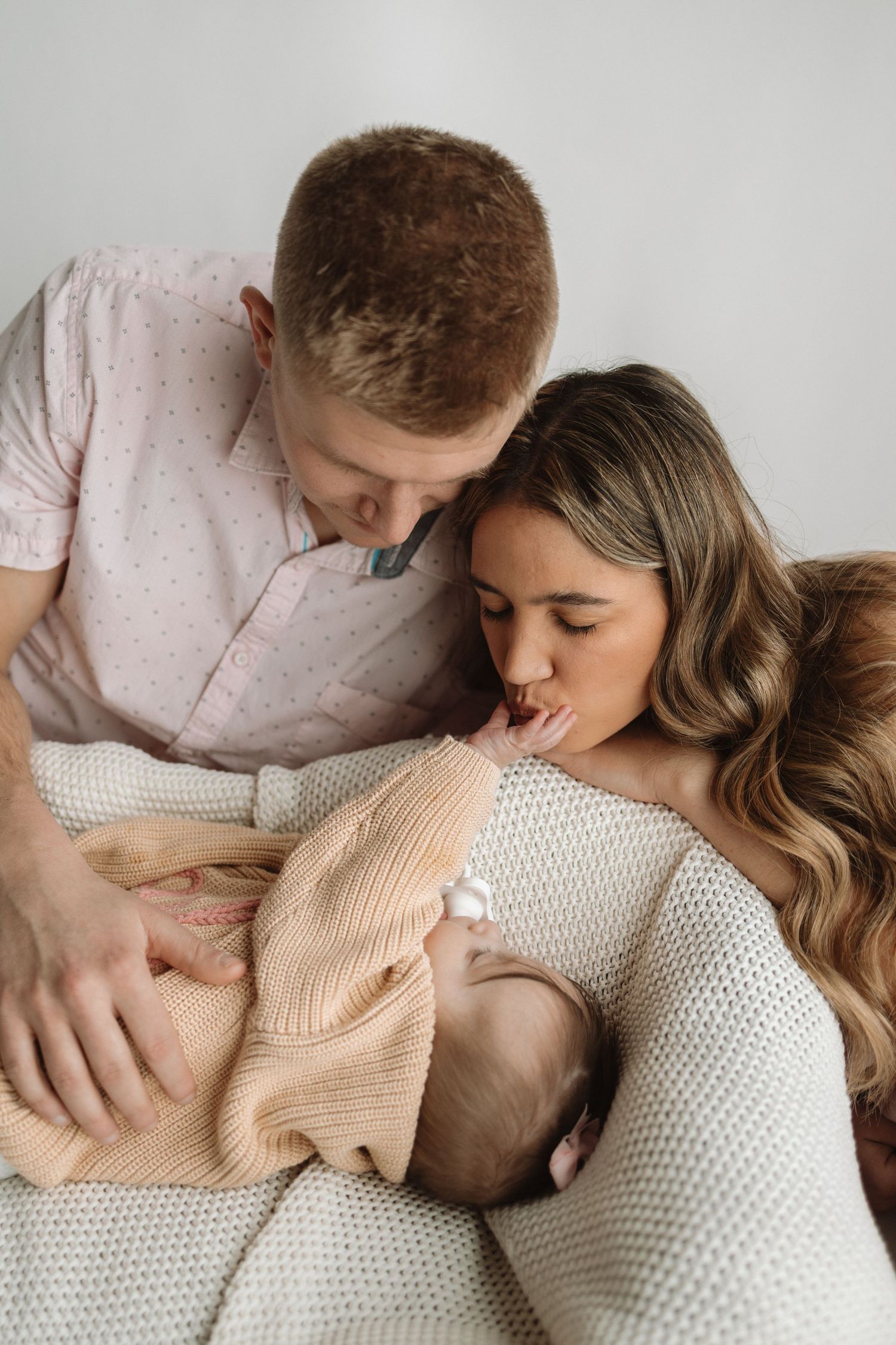 Intimate family moment showing parents cuddling with newborn baby on white textured bedding.