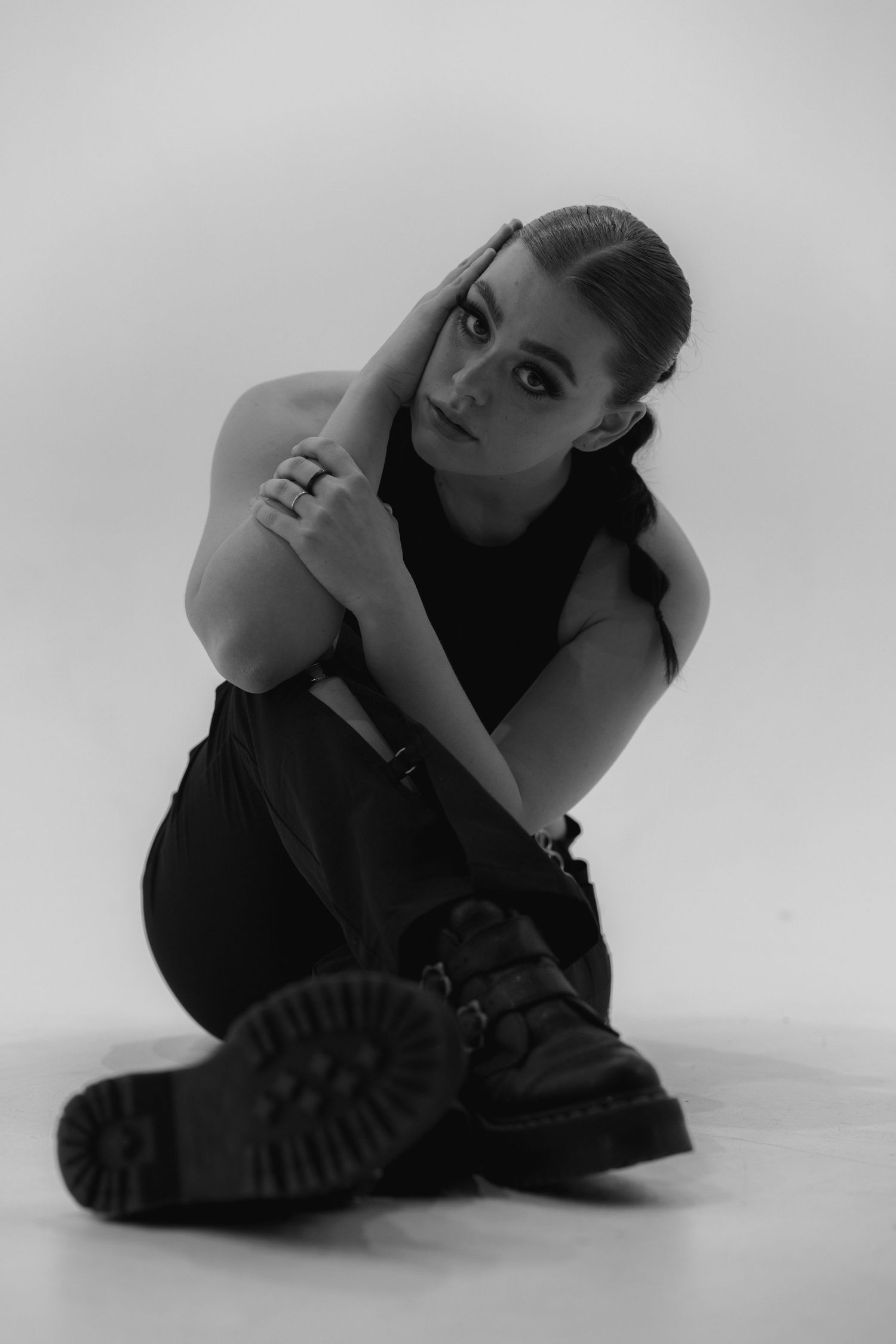 Black and white portrait of figure in dark outfit and combat boots sitting on white floor in artistic pose.