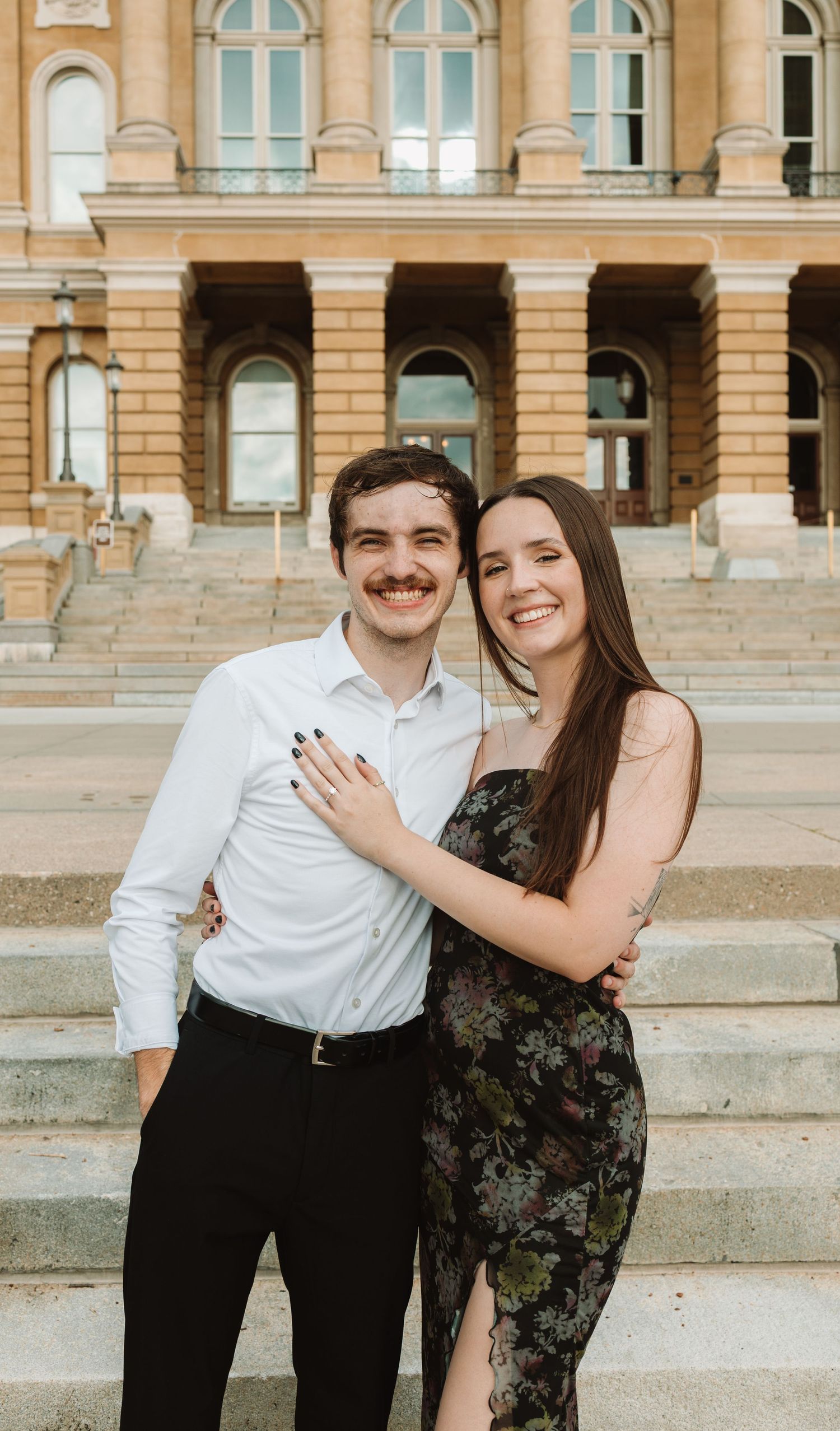 A couple stands together on stone steps outside a grand historic building with arched windows and ornate architecture.