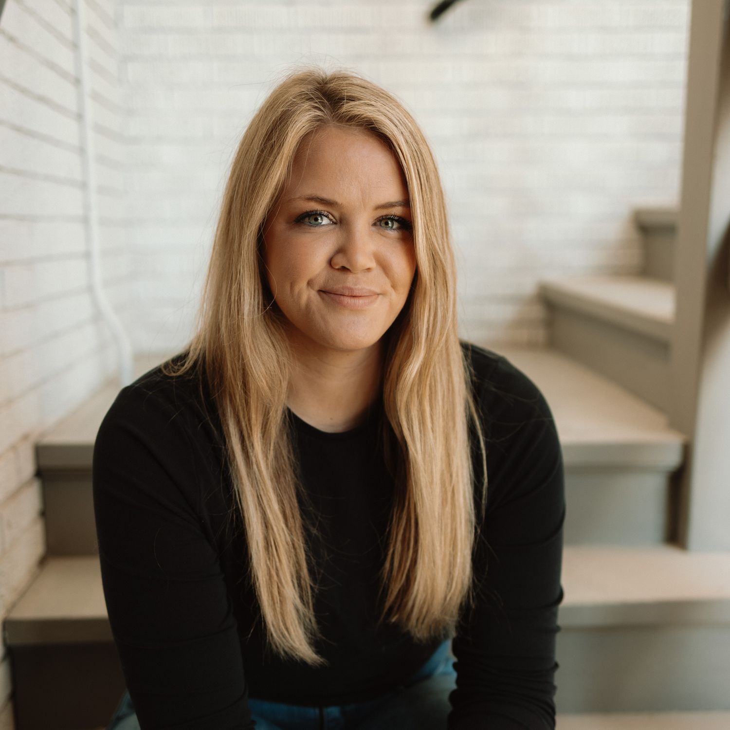 Young woman with long blonde hair wearing a black sweater sits on stairs with a warm smile.