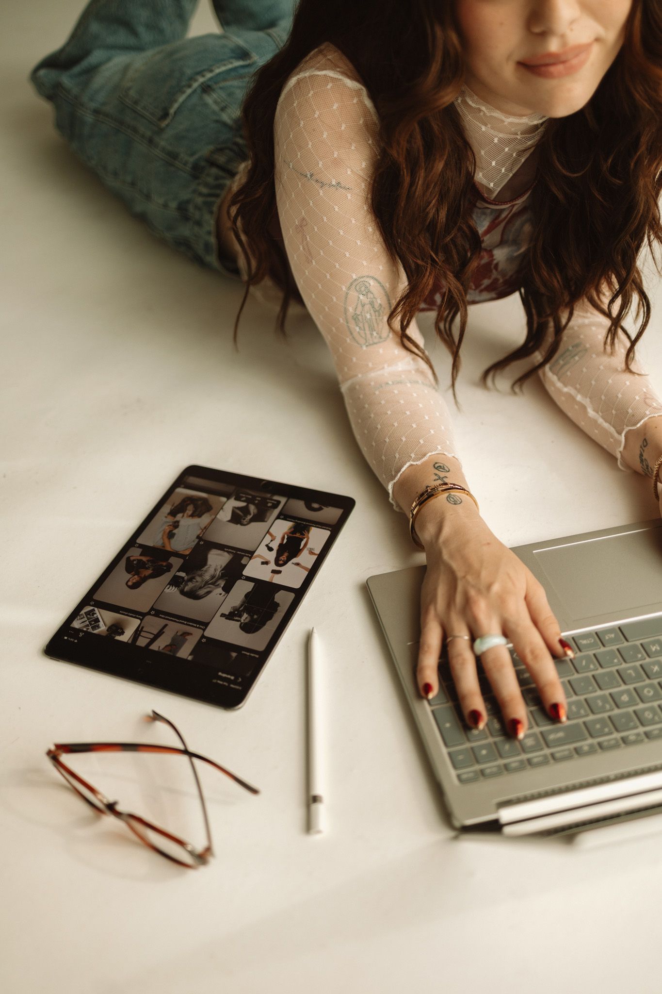 Person in lace top working on laptop with tablet and glasses on white surface.
