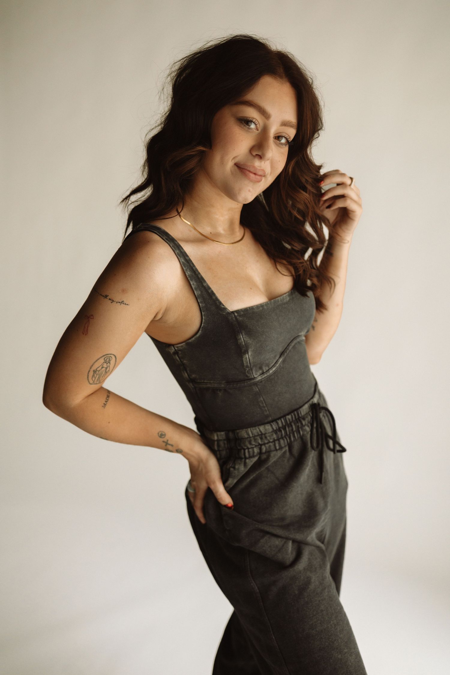 Woman in stylish dark green overalls poses against light background showing visible arm tattoos and wavy dark hair.