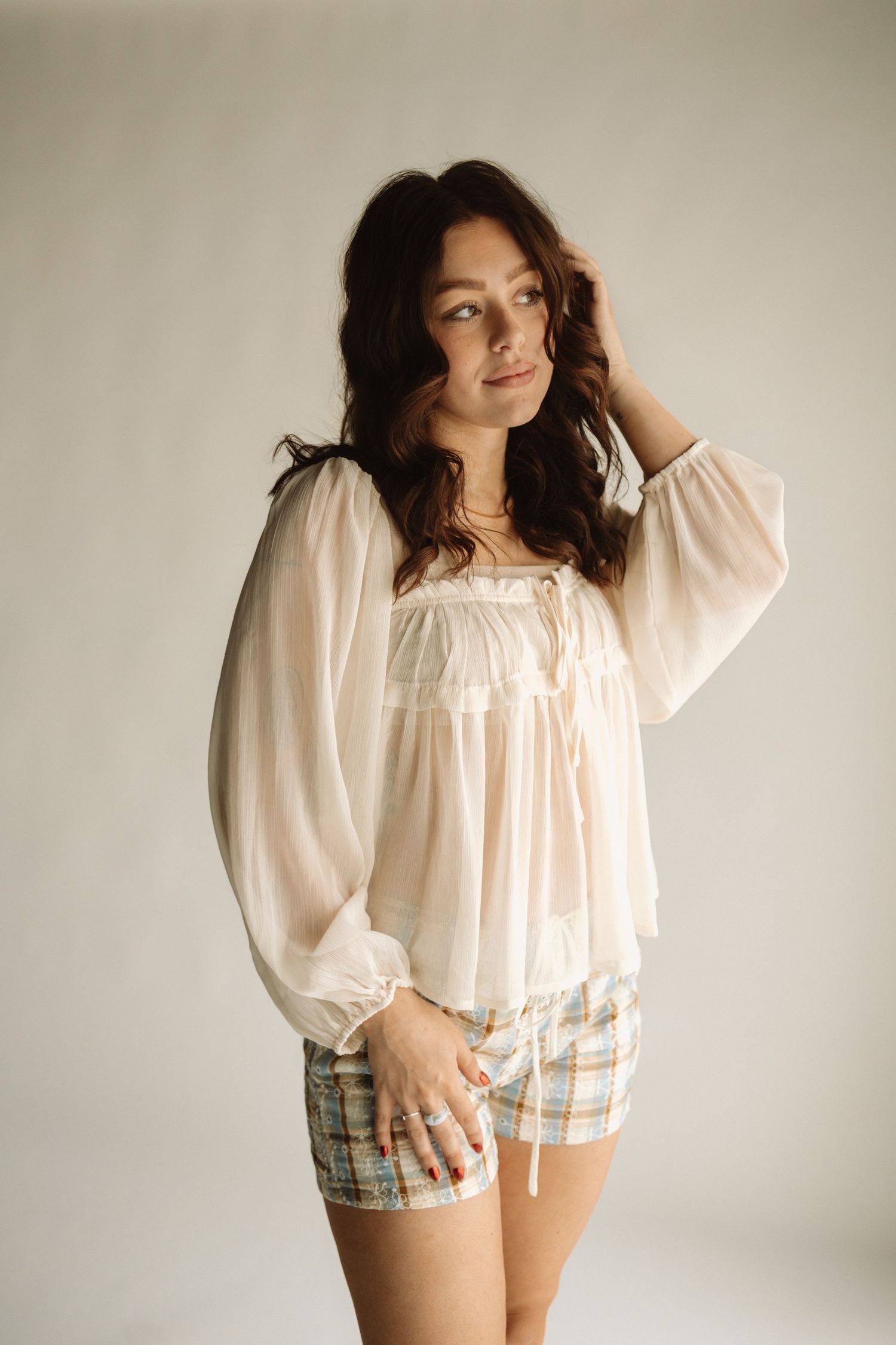 A woman poses in a romantic white peasant blouse and plaid shorts against a neutral backdrop.