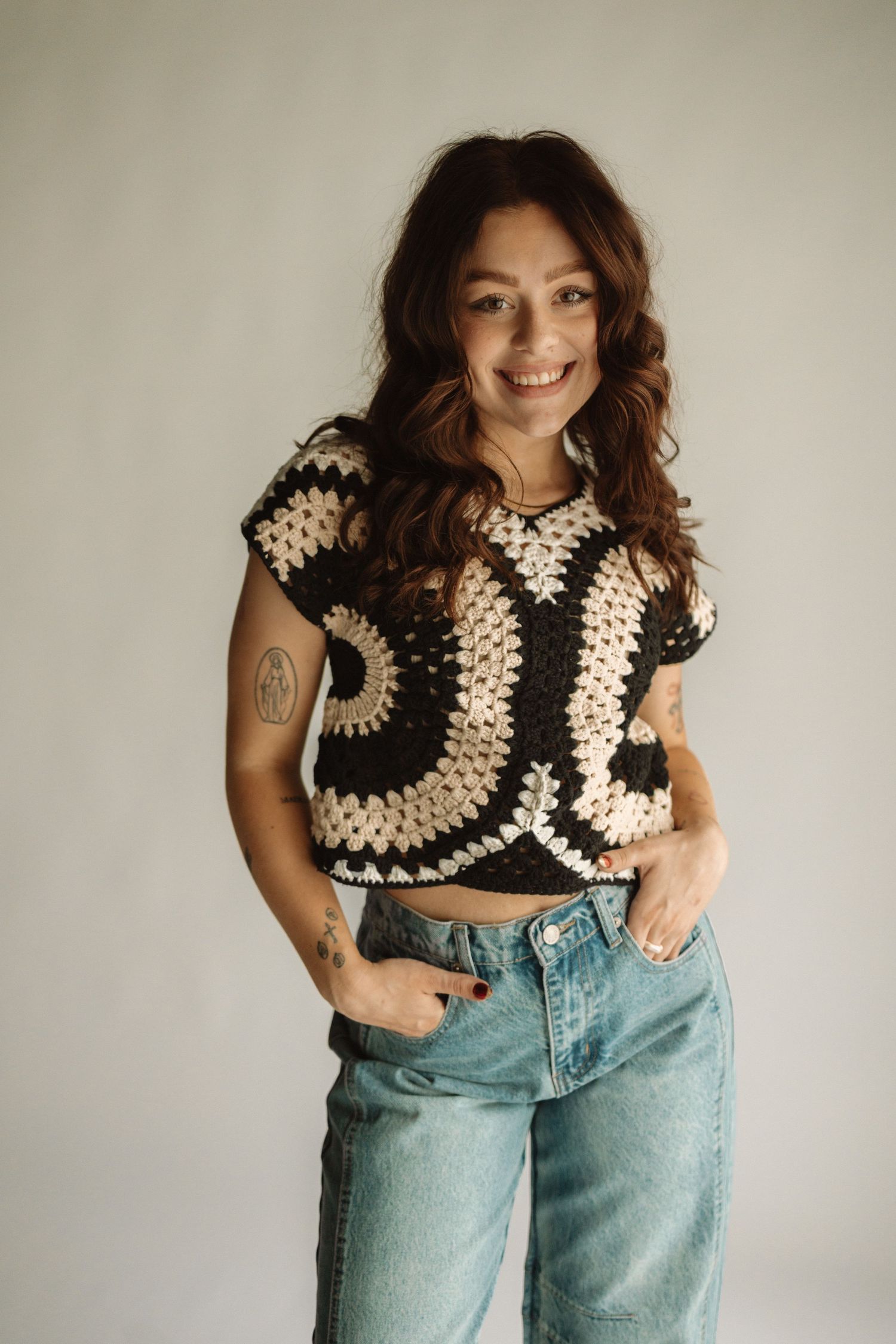 A person wearing a black and cream crochet crop top and light wash jeans poses against a neutral wall.