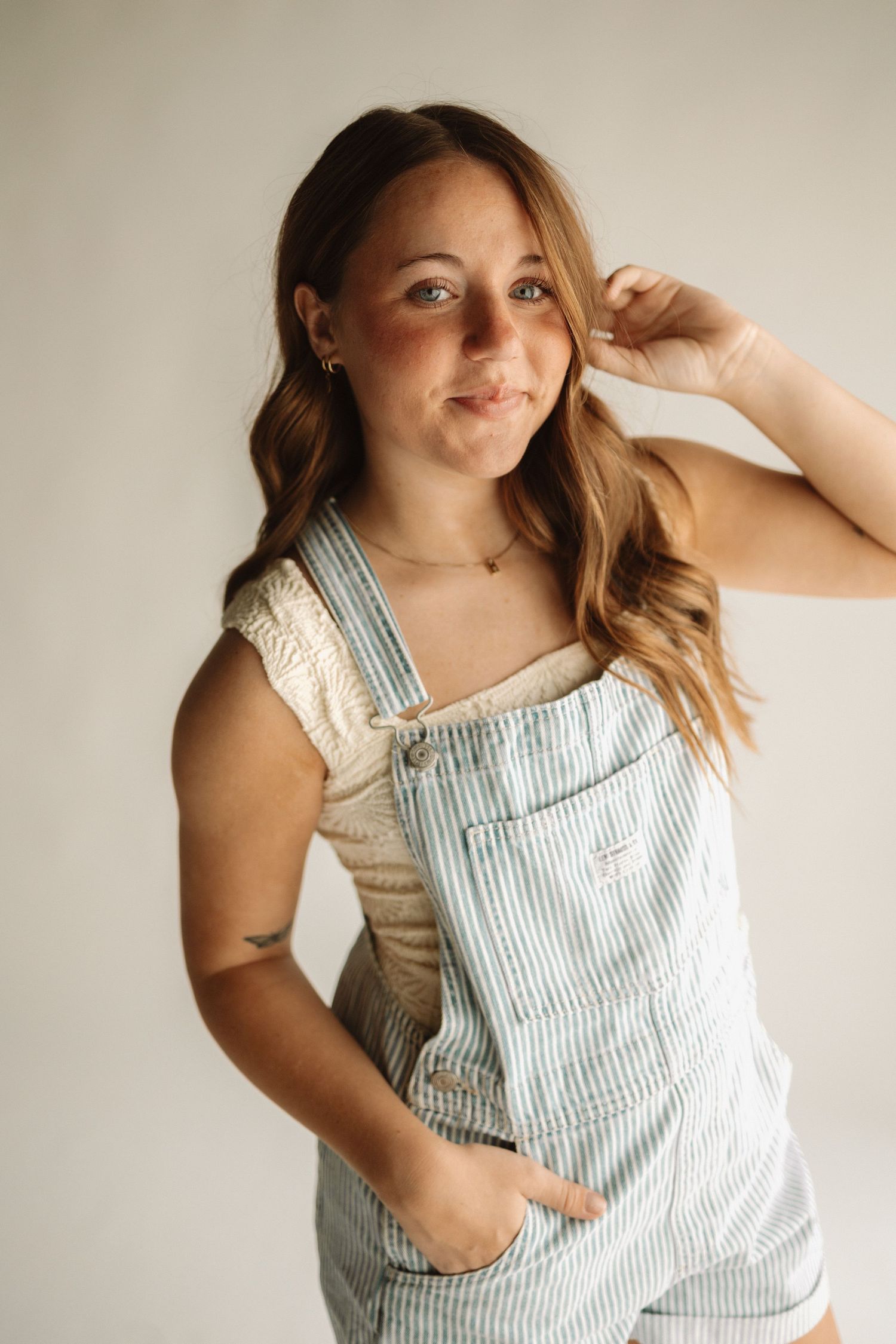 Woman with wavy brown hair poses casually in light blue and white striped overalls against neutral background.
