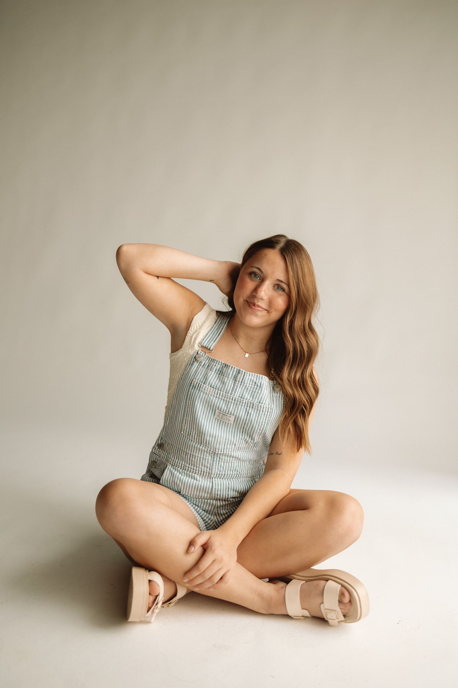 A person in light blue overalls and sandals sits cross-legged on a white backdrop with natural lighting.
