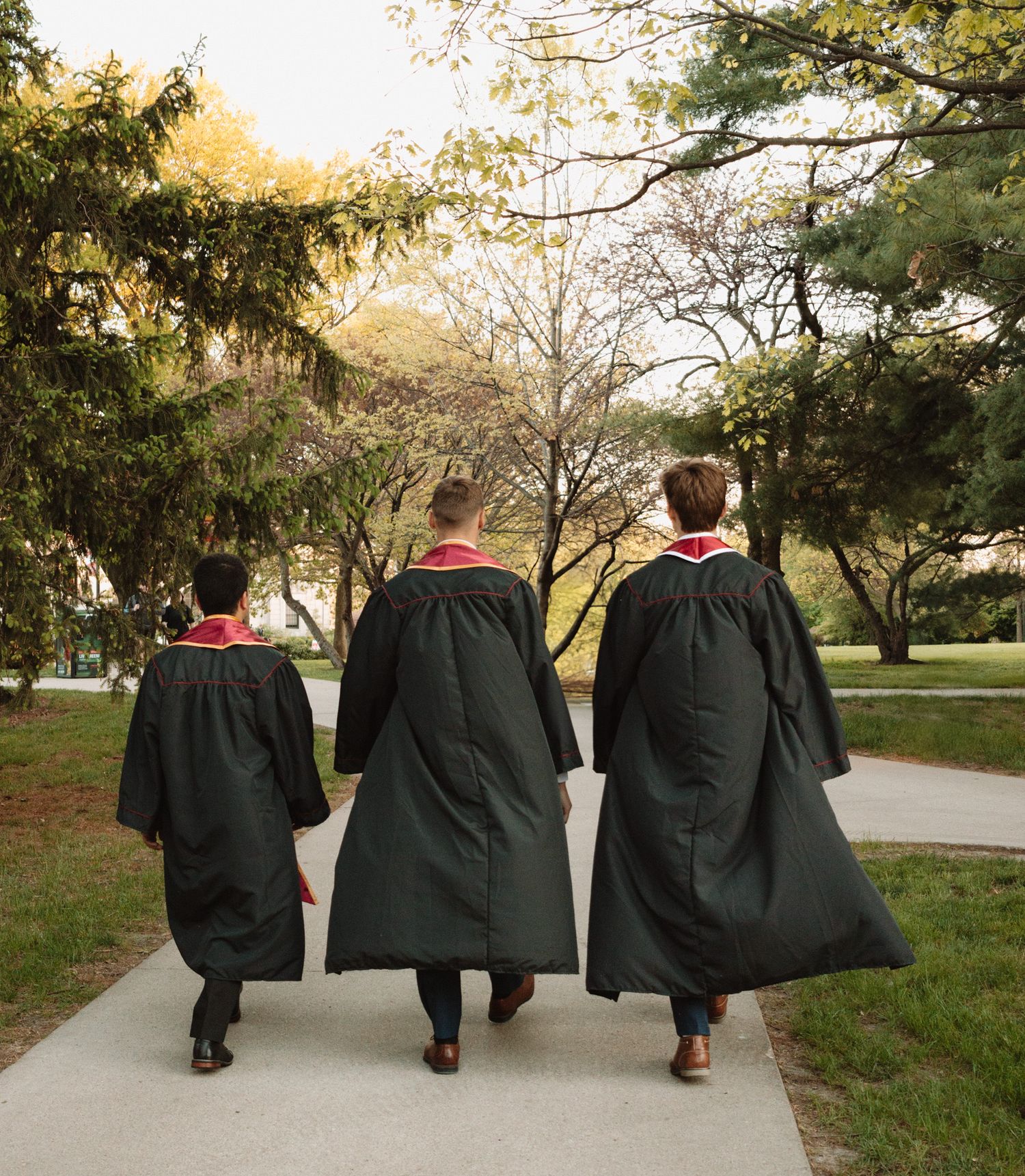 Three graduates in black graduation gowns walk together on a campus path at sunset.