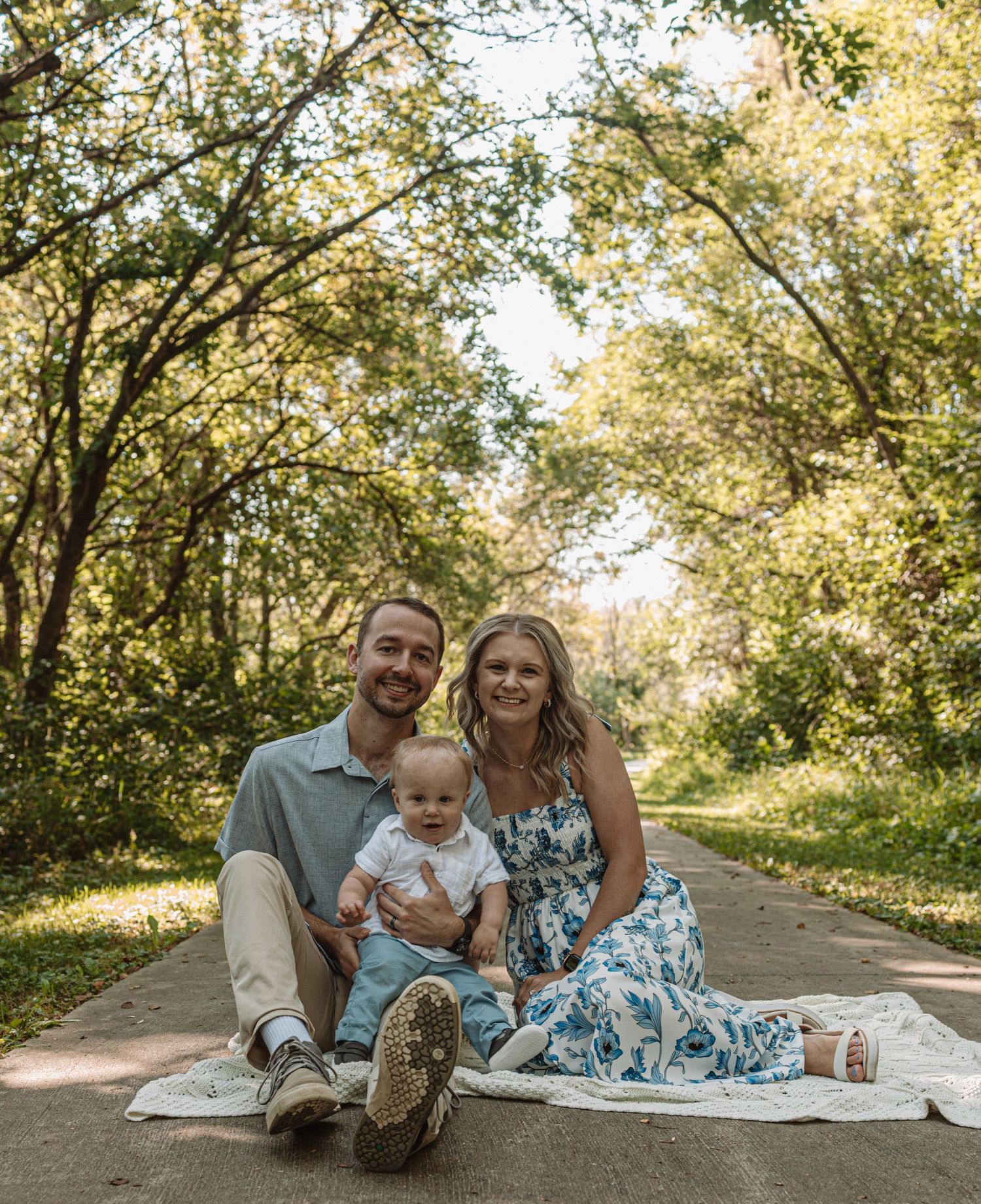 A family relaxes on a white blanket under sun-dappled trees along a peaceful walking path on a warm day.