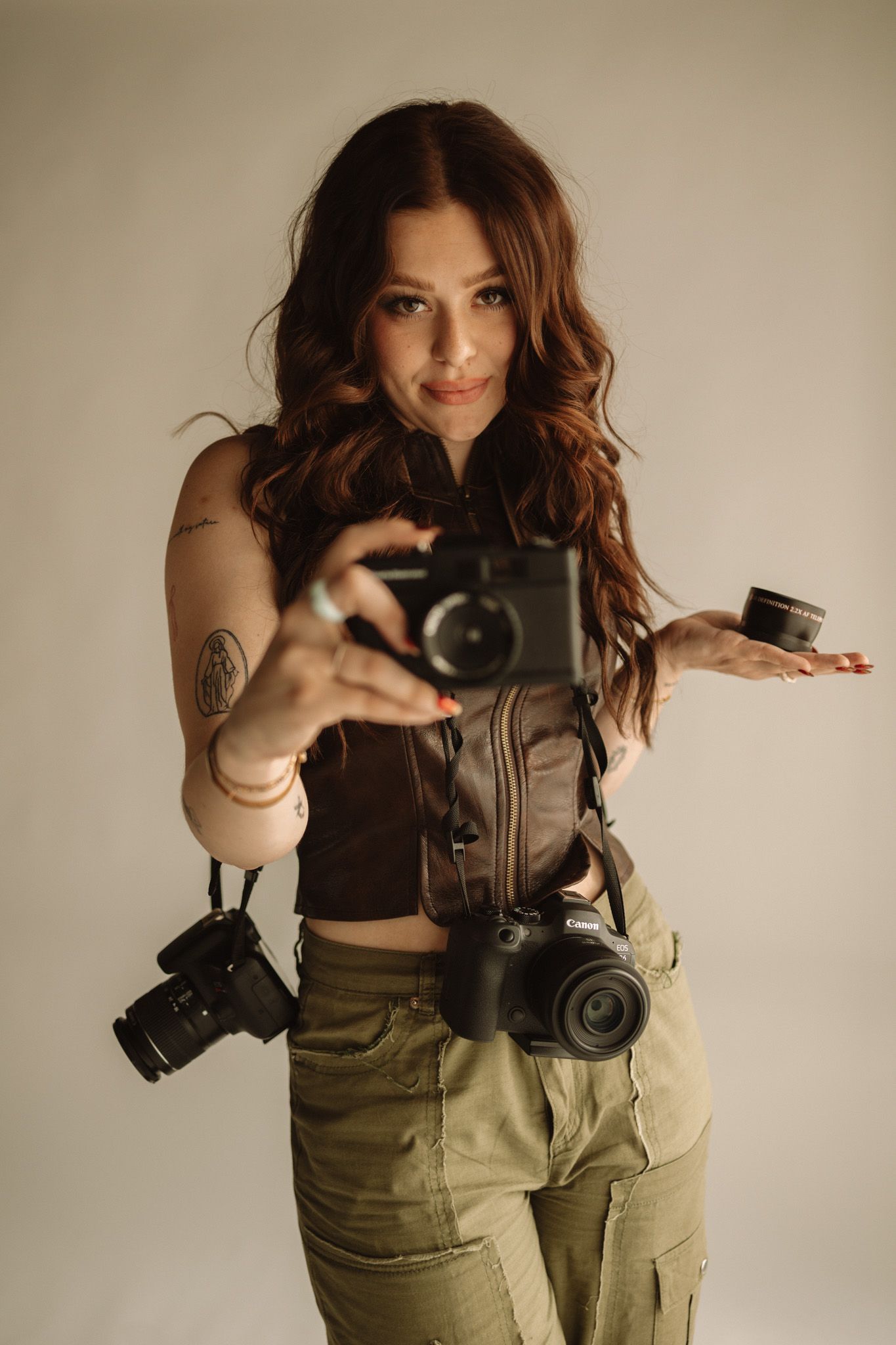 Creative photographer poses with multiple cameras and cargo pants in moody studio portrait.