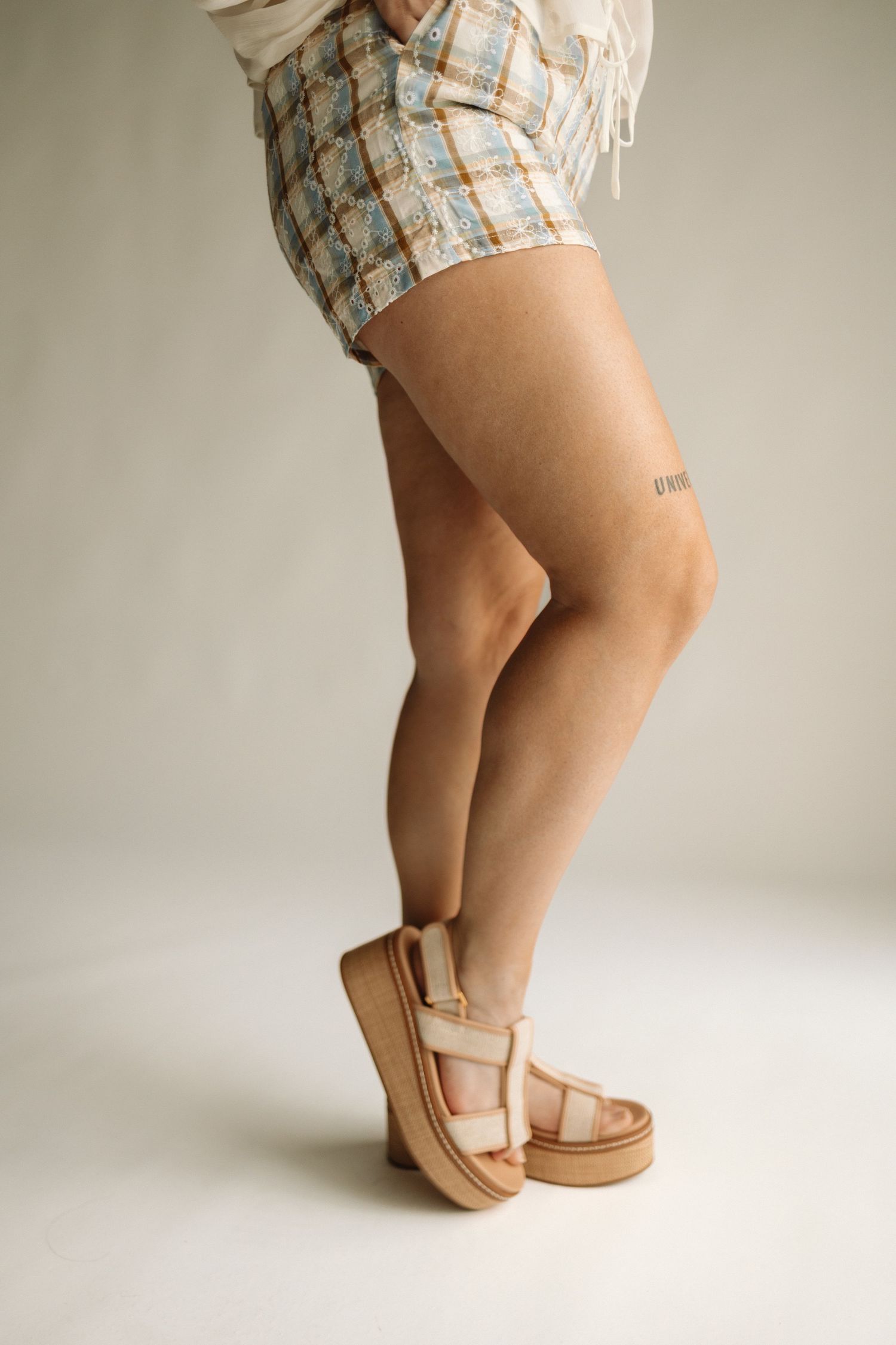 Legs wearing plaid shorts and beige platform sandals stand against neutral background in summer fashion pose.