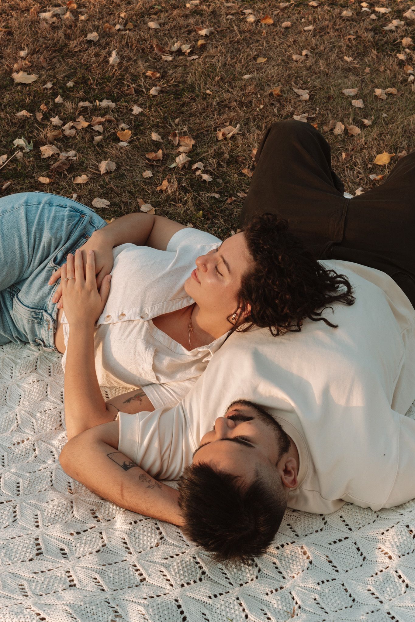 Two people laying together on a white lace blanket outdoors among fallen autumn leaves.