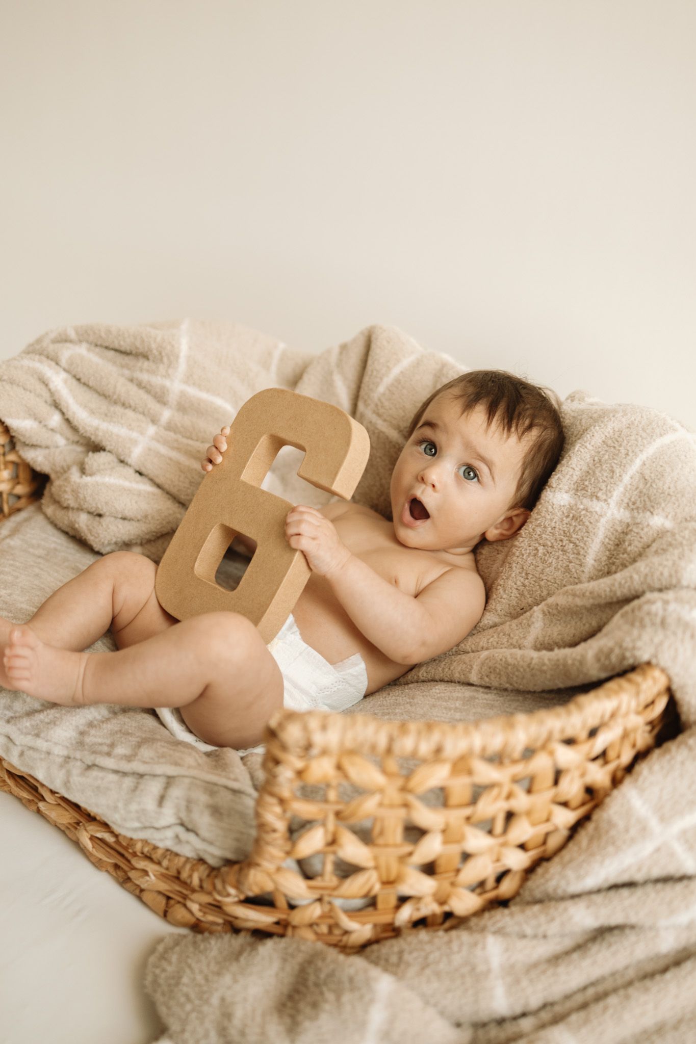 Baby in diaper holding wooden number 6 while laying in woven basket with beige blanket.
