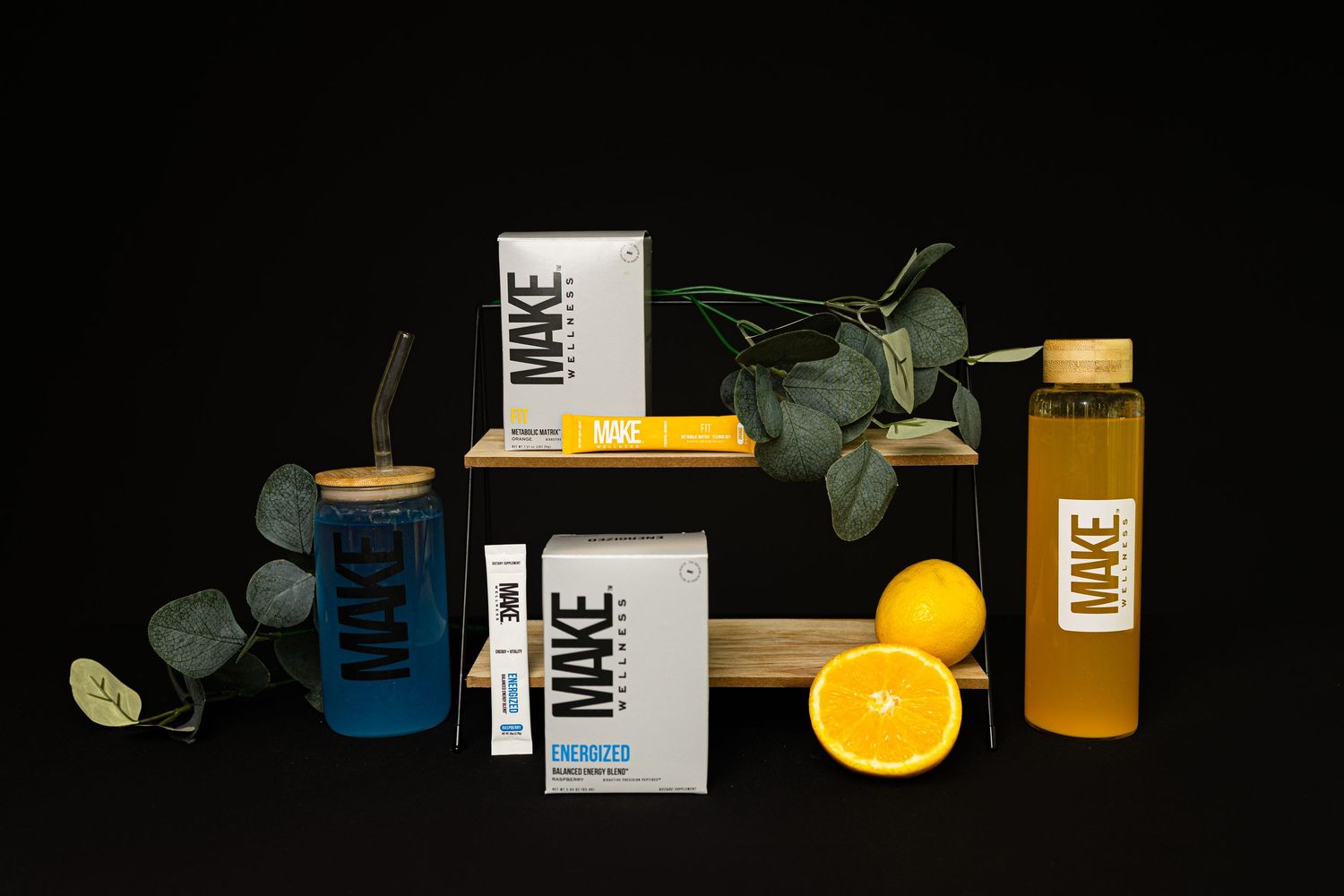 Assorted juice bottles and product packaging arranged with eucalyptus leaves and fresh orange slices on black background.