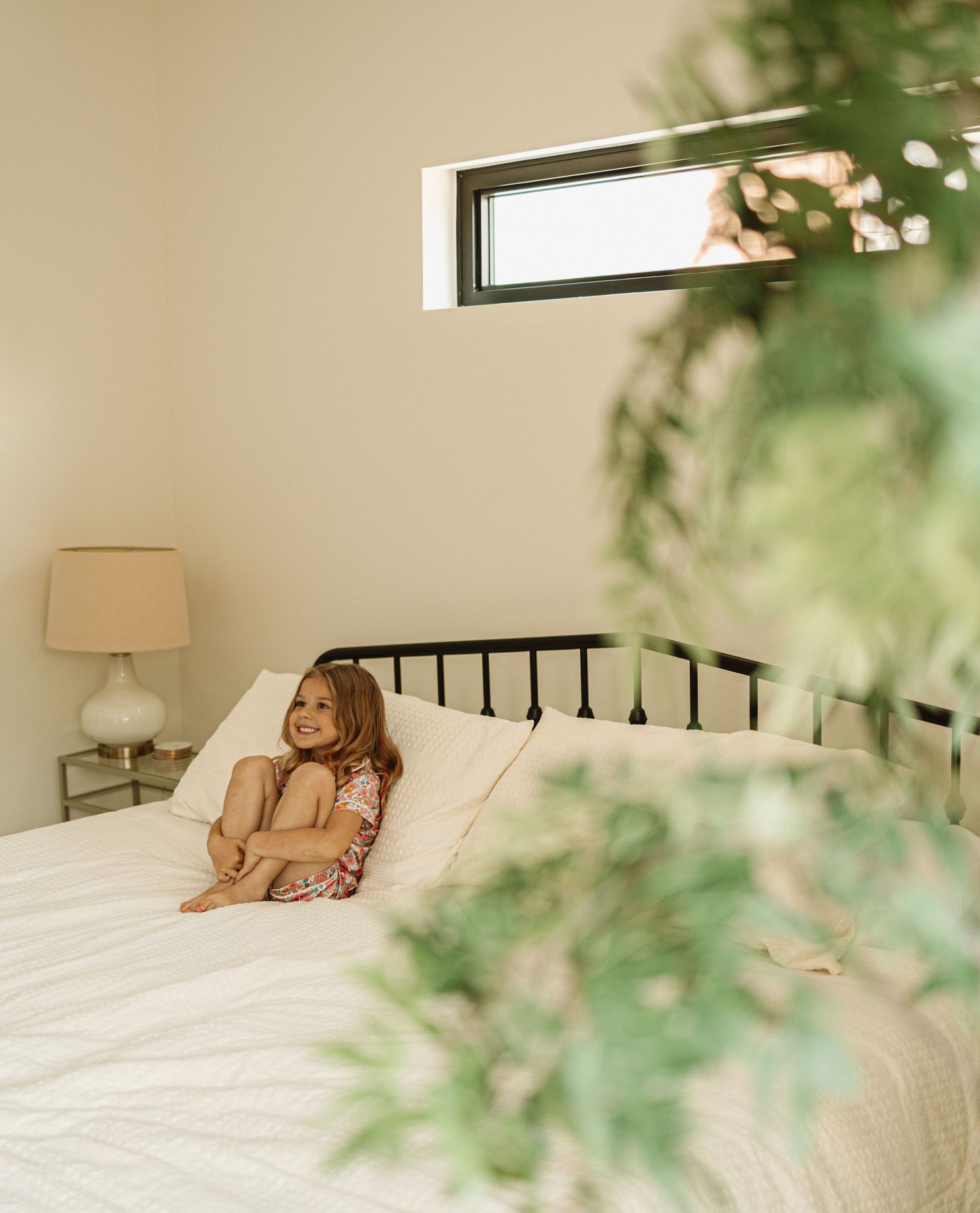 A cozy bedroom with a bed, lamp, and tranquil natural light filtering through a window, framed by green plants.