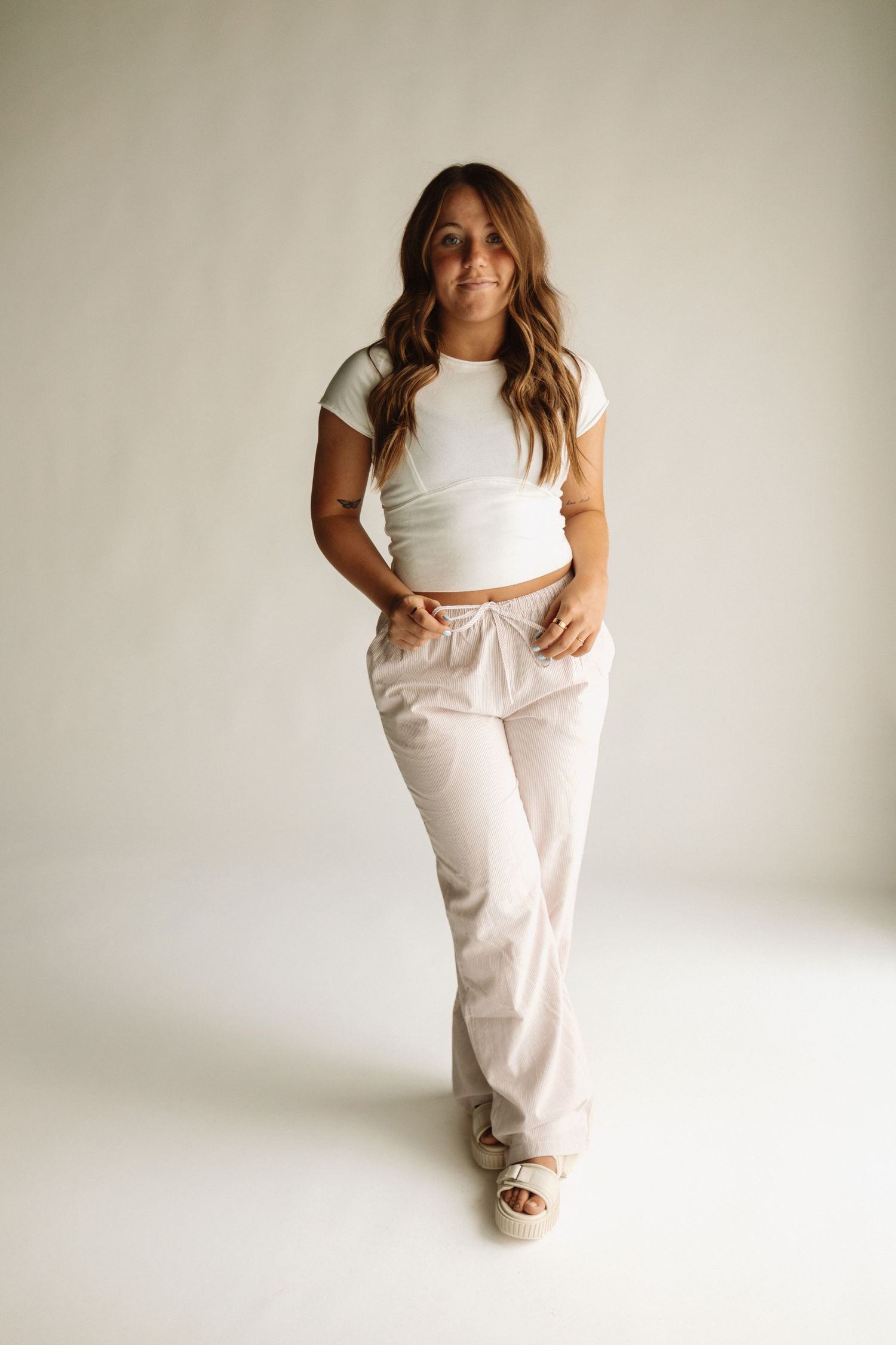 Someone poses in a casual white crop top and beige linen pants against a neutral backdrop, wearing platform sandals.