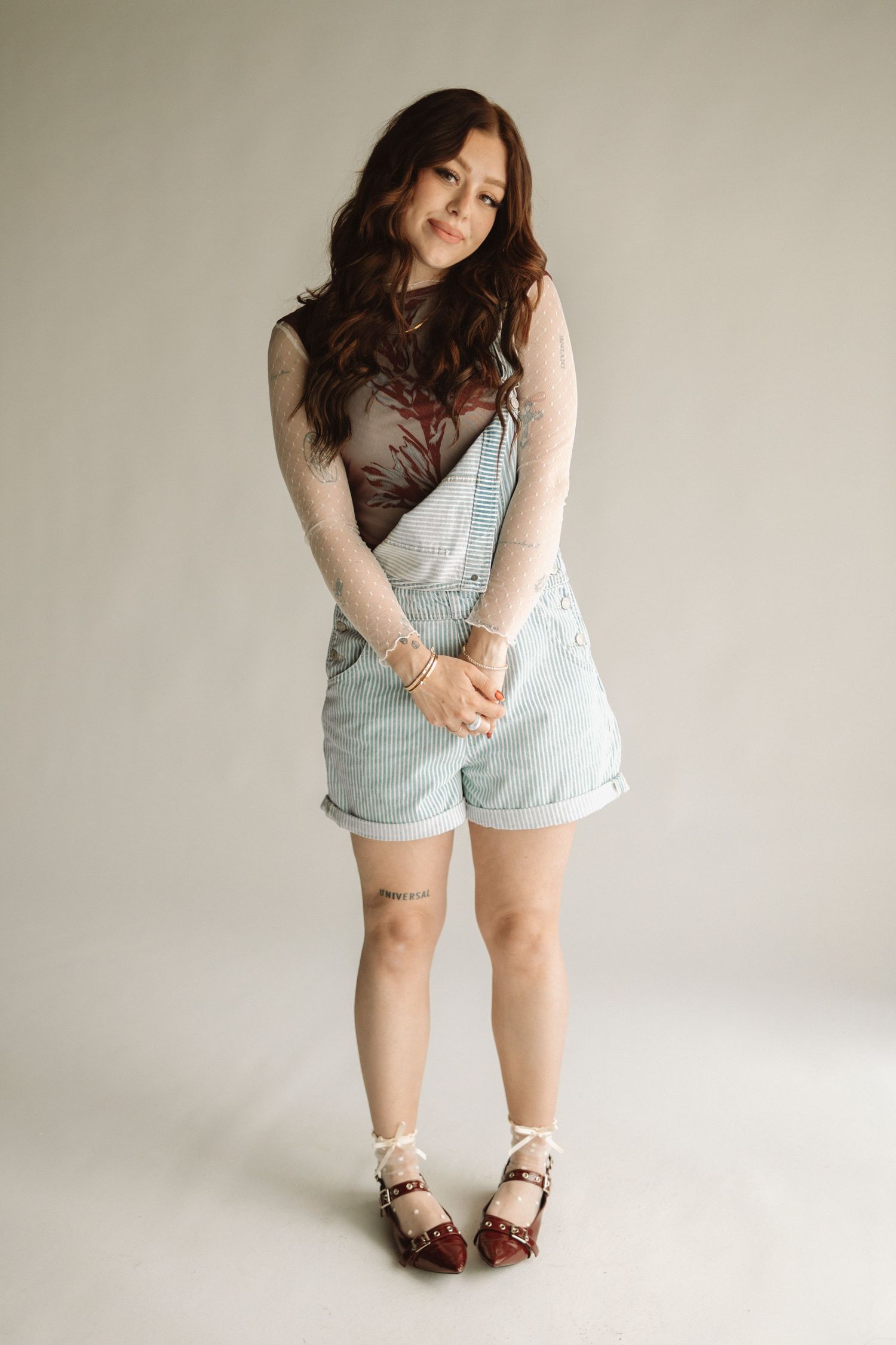 A person wearing light denim overalls and a sheer long sleeve top poses casually against a neutral background.