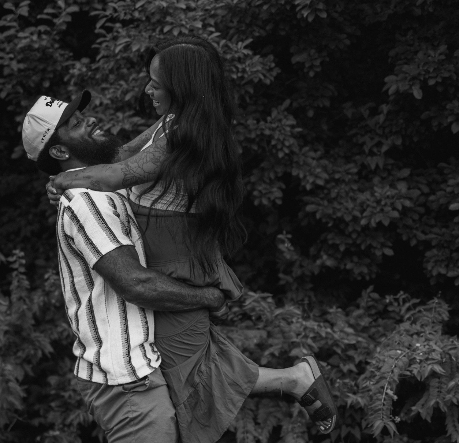 Black and white photograph of two seated figures embracing outdoors among foliage in a romantic pose.