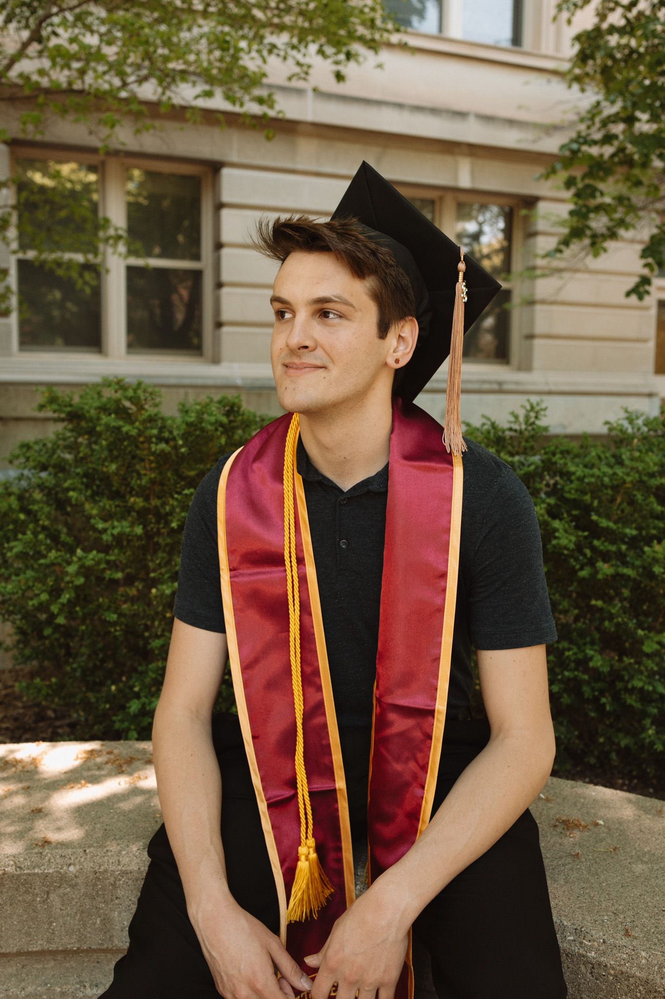 Graduate in black cap and burgundy graduation sash poses outdoors in front of stone building on sunny day.