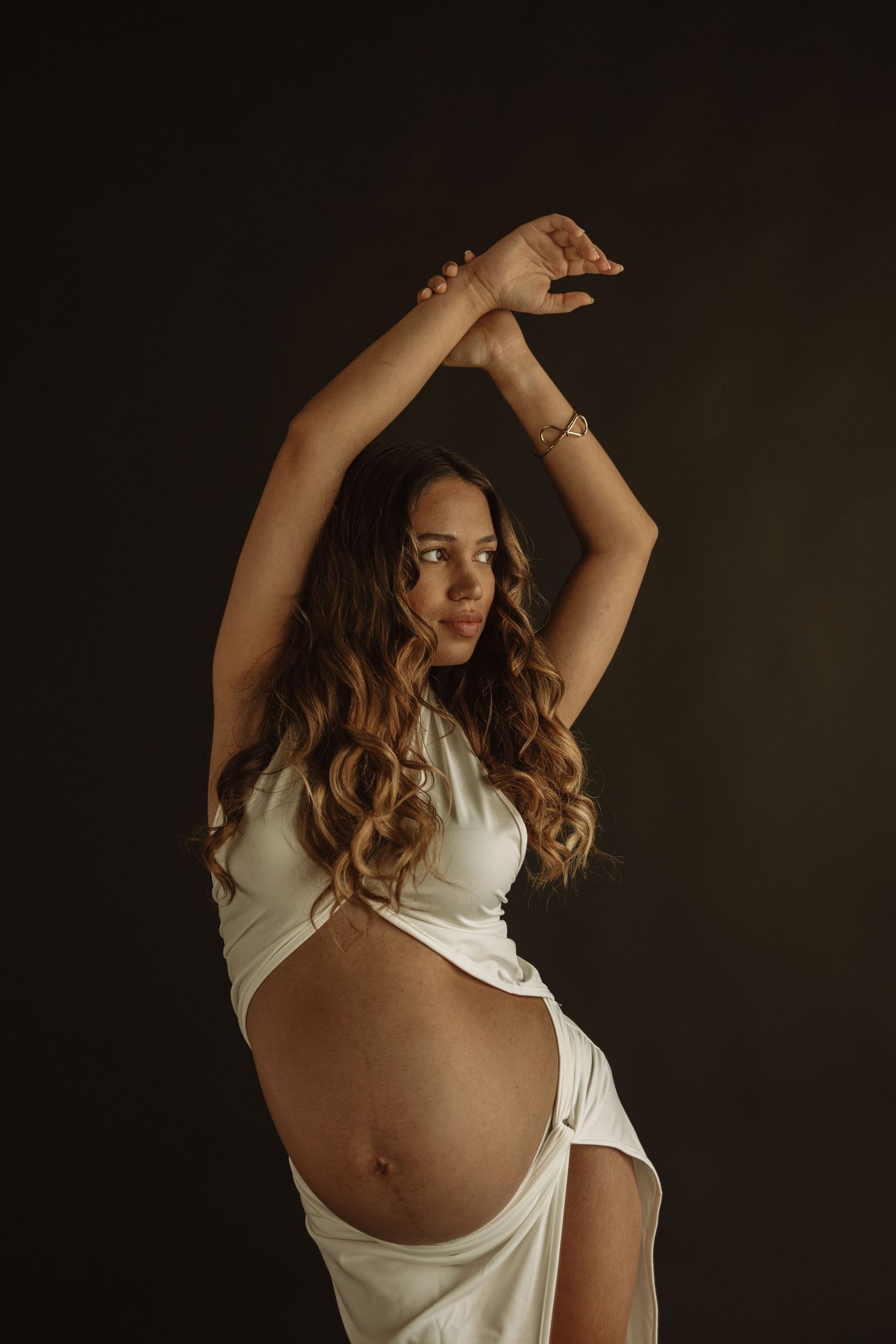 Artistic maternity portrait features an expectant mother in white fabric posing elegantly against a dark background.