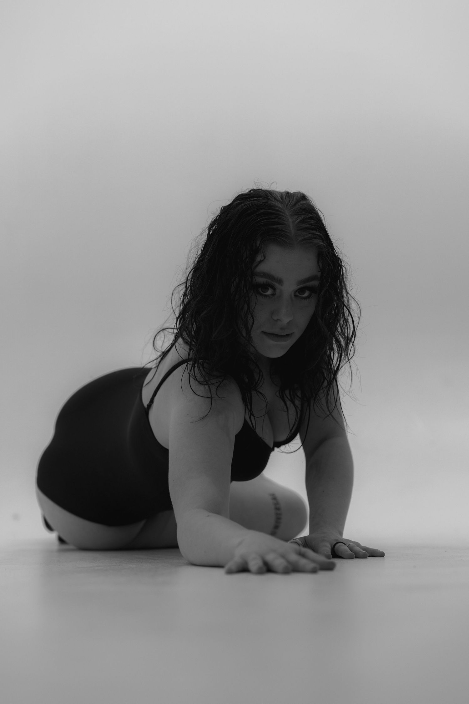 Black and white artistic portrait photo of someone posing on the floor in dark attire with wet-looking hair.