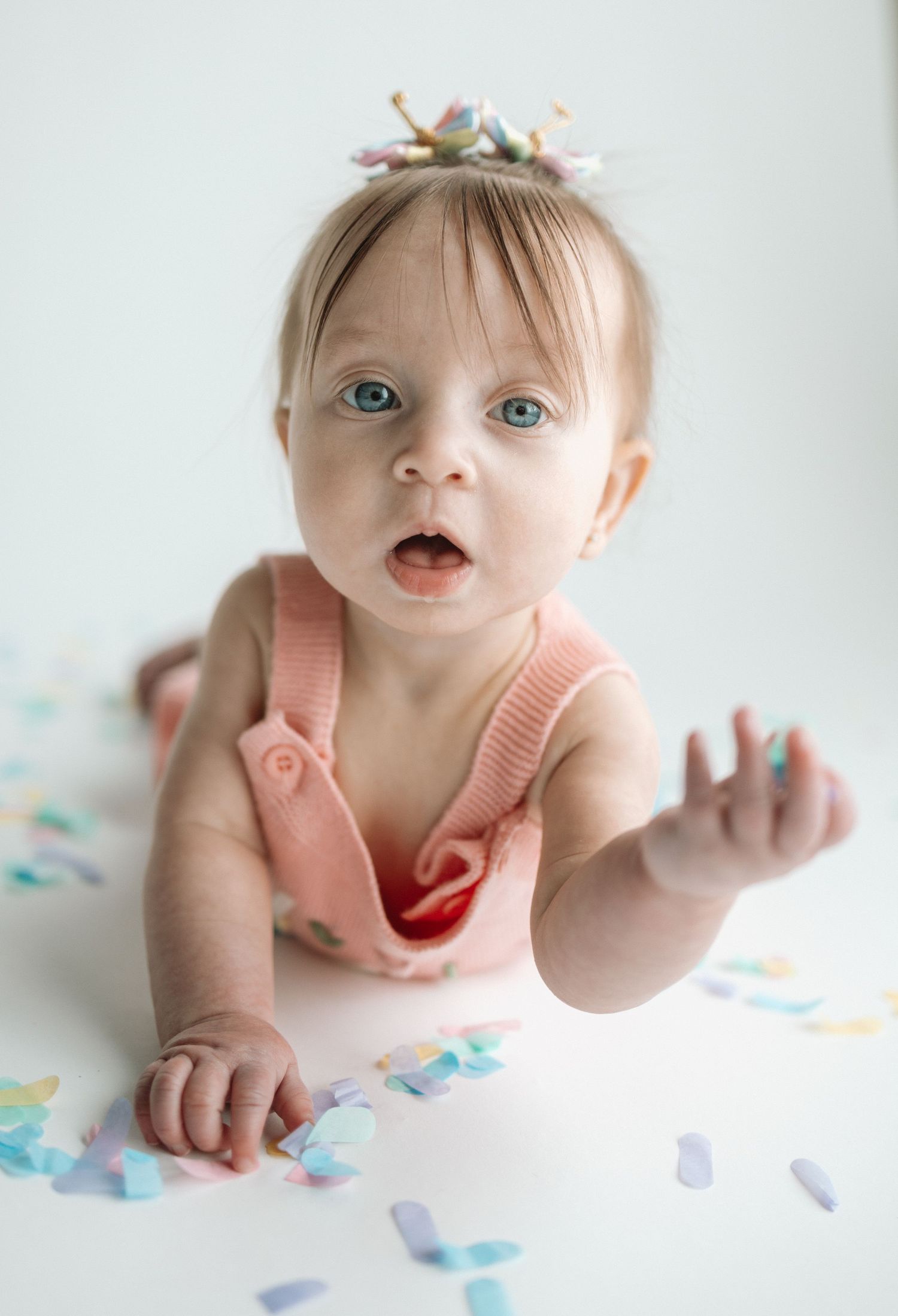 Baby wearing a peach-colored outfit lies on white surface surrounded by pastel paper confetti.