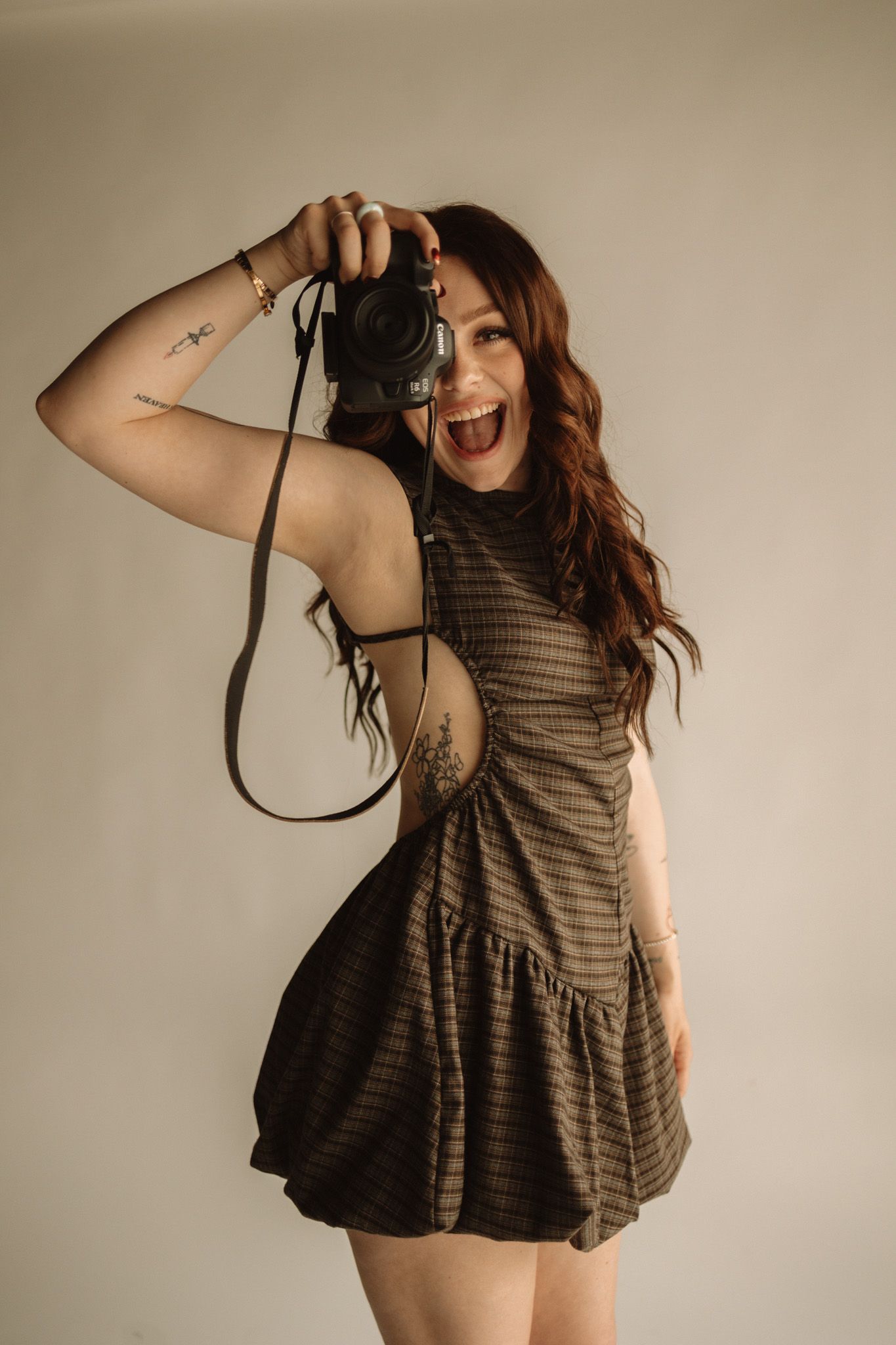 A photographer wearing a brown striped sundress holds up a vintage camera while expressing joy and excitement.
