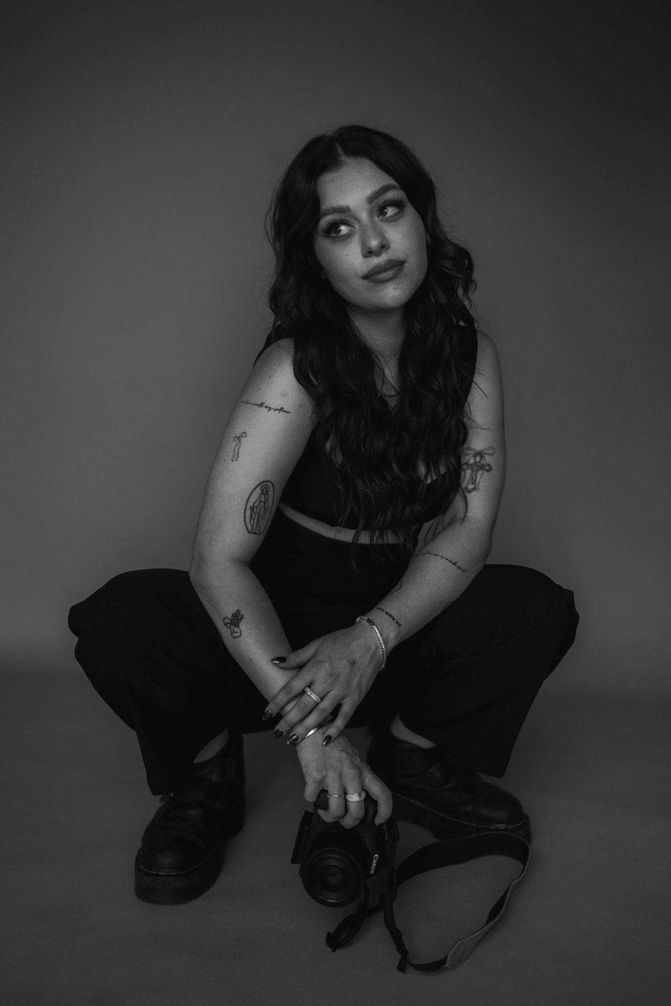 Black and white portrait of a photographer sitting casually in dark clothing with visible tattoos in a studio setting.