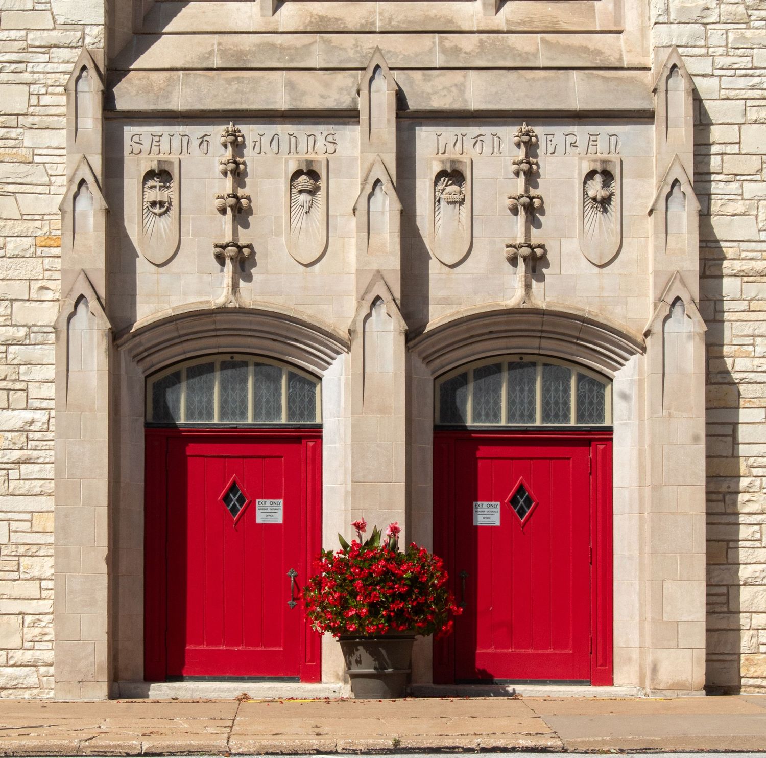 Two bright red church doors with arched stone surroundings and traditional lantern lights mounted on a limestone facade.
