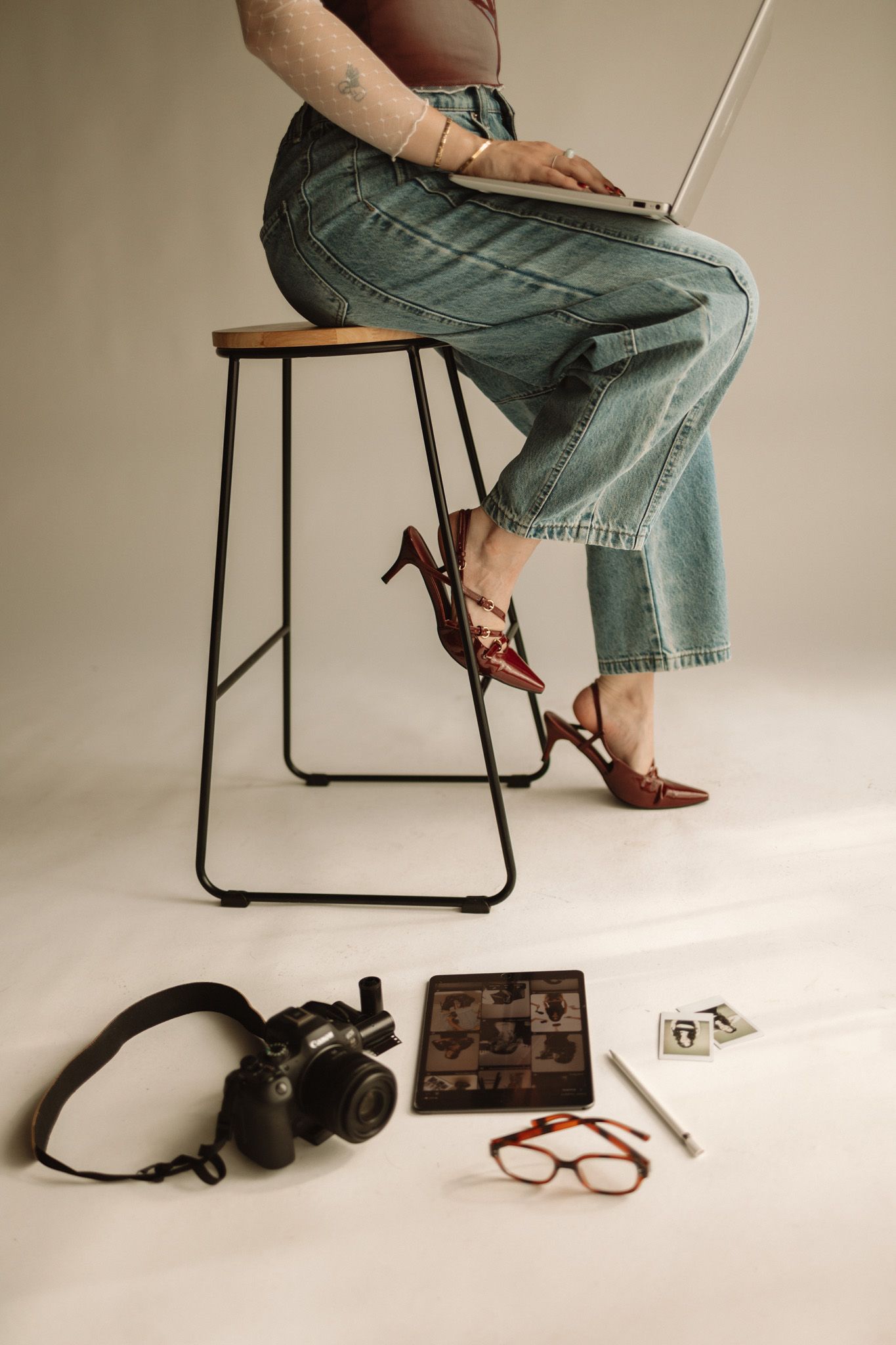 A person sits on a modern stool wearing jeans and brown shoes, with photography equipment and glasses arranged below on white floor.