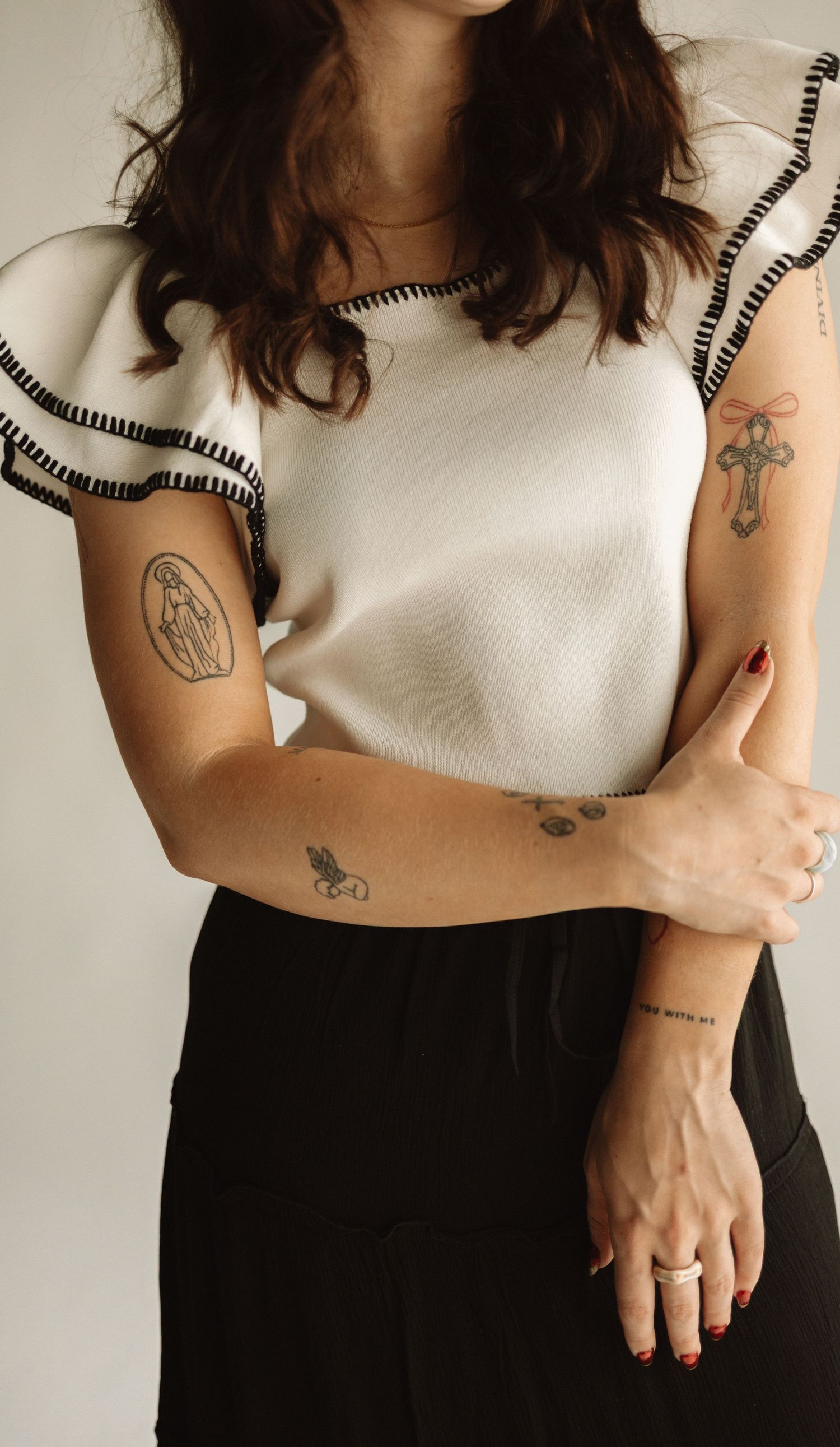 Person wearing white sleeveless top with black trim and dark pants showing arm tattoos against light background.