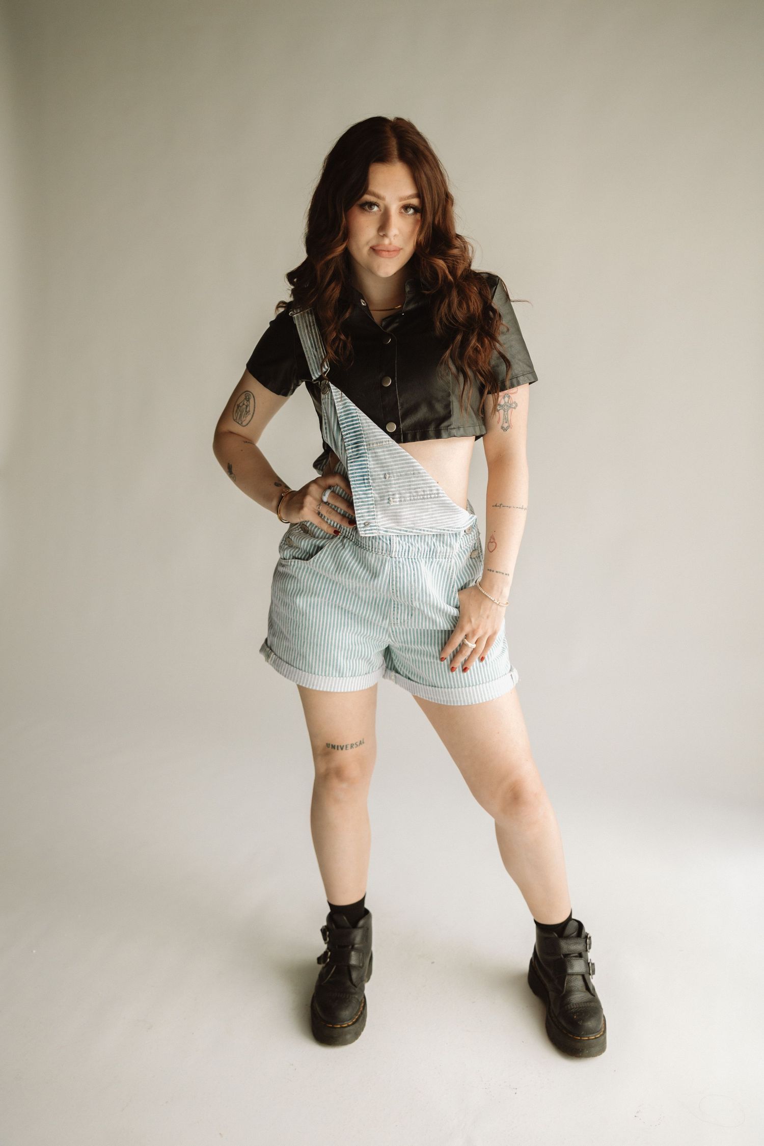 Small child sits on floor in overalls and black boots against white wall and poses in different positions.