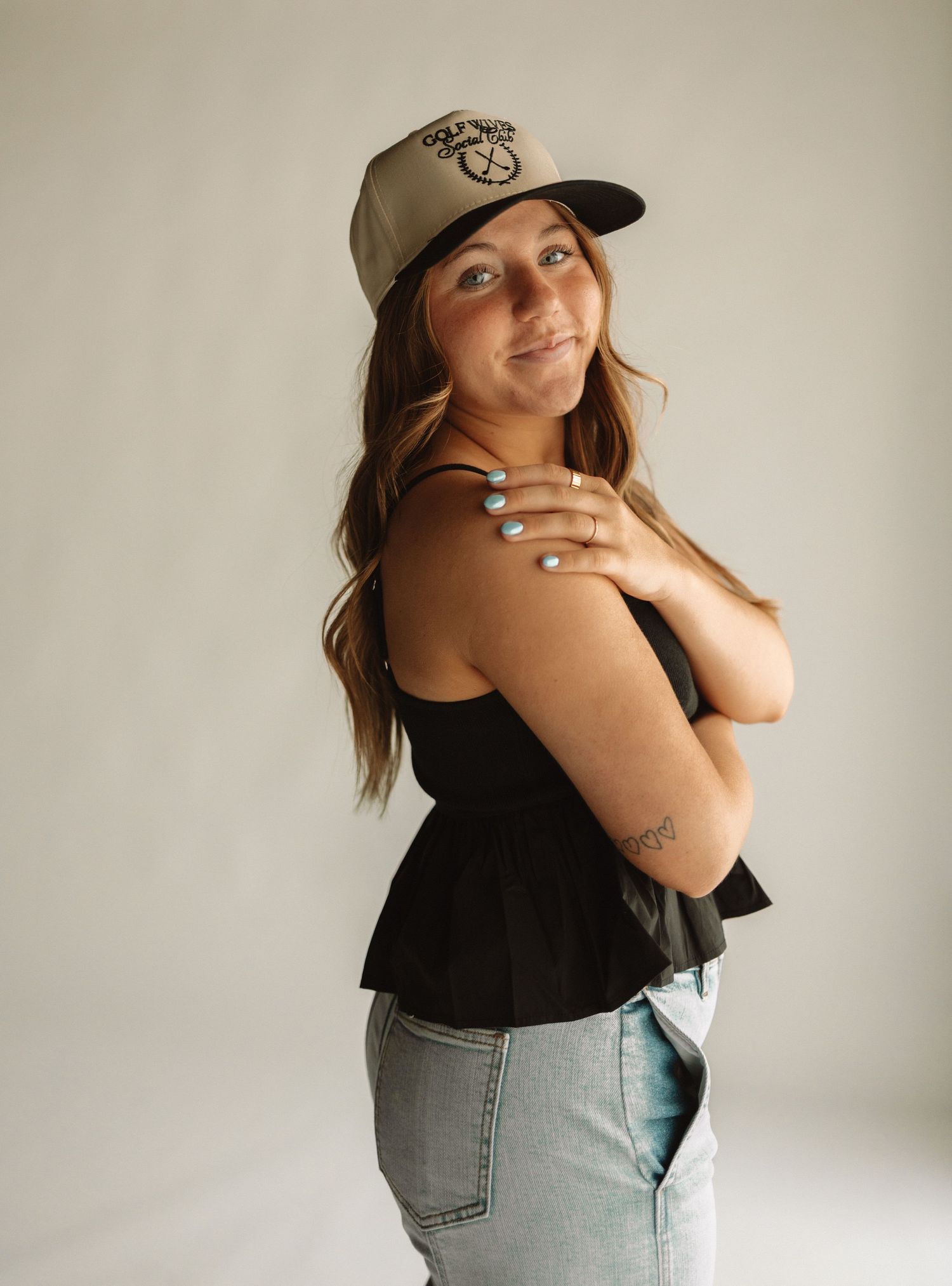 Person wearing a baseball cap and black sleeveless top poses against white background with arms crossed.