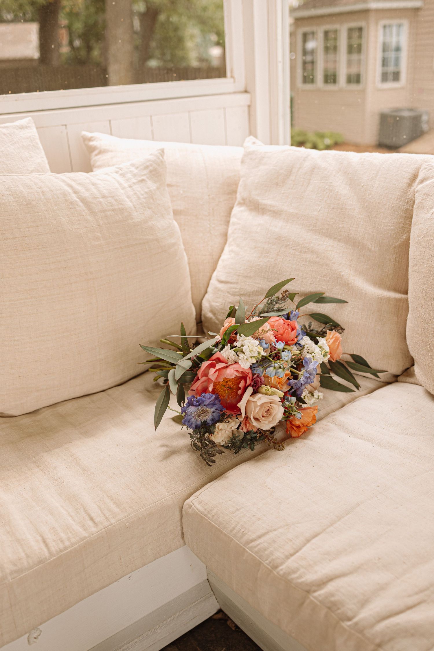 A colorful floral arrangement with peach roses and blue blooms rests on a plush cream-colored couch cushion.