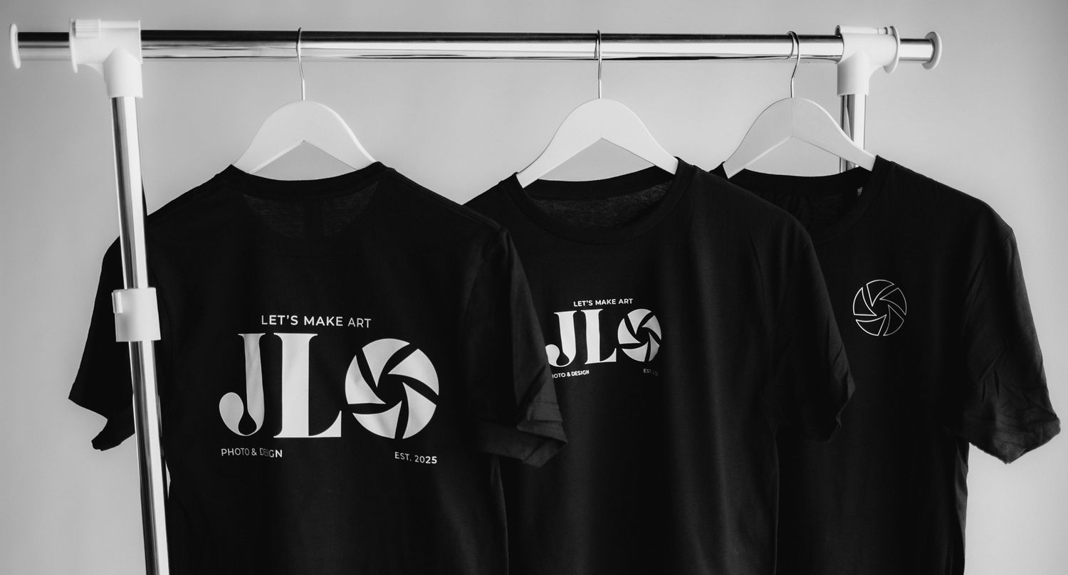 Three black t-shirts with JLS branding hanging on white clothing rack.